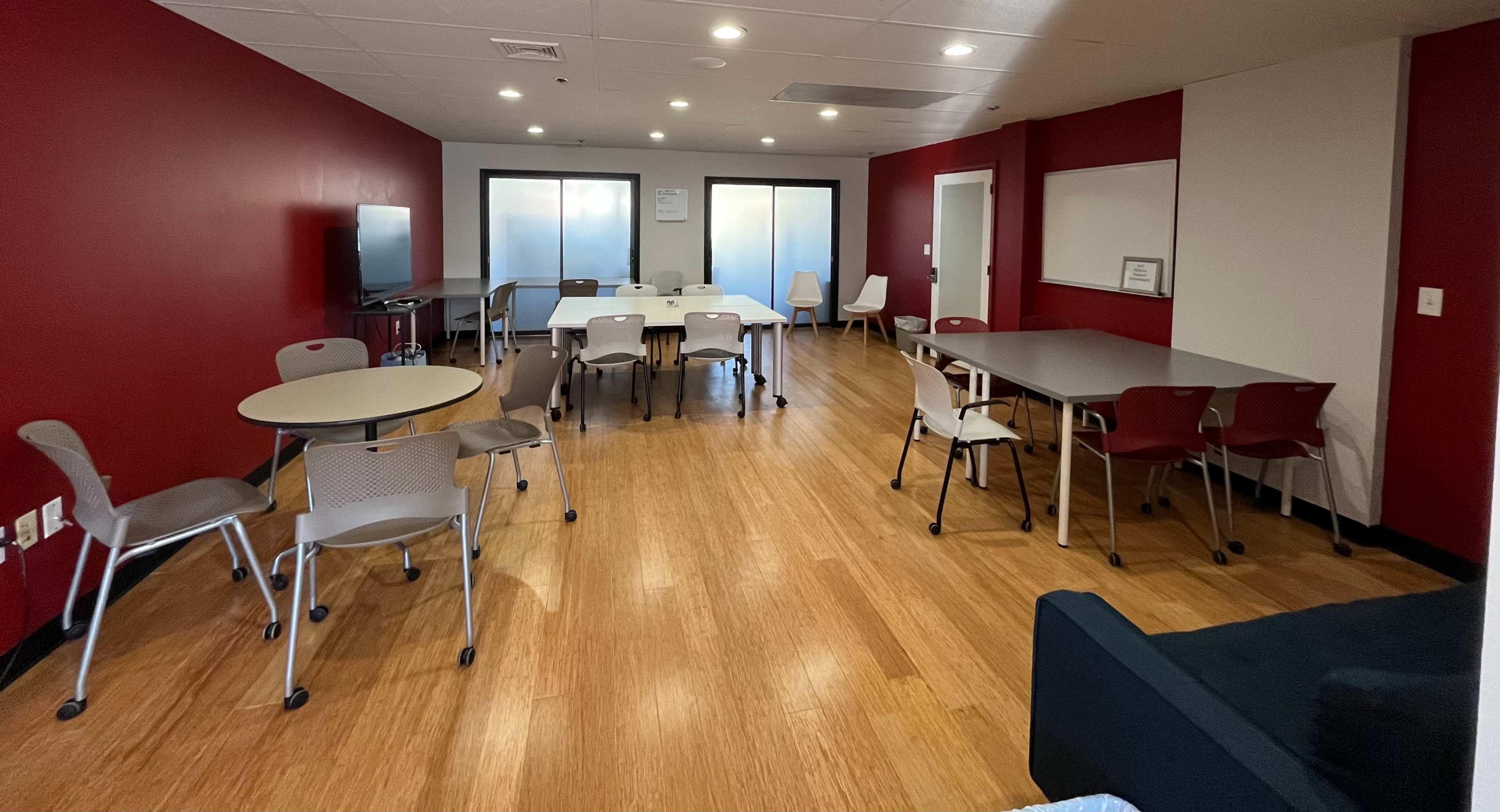 The image shows a communal room with several tables and chairs, a TV, and a whiteboard on the walls, all set on a bamboo floor with a red accent wall.
