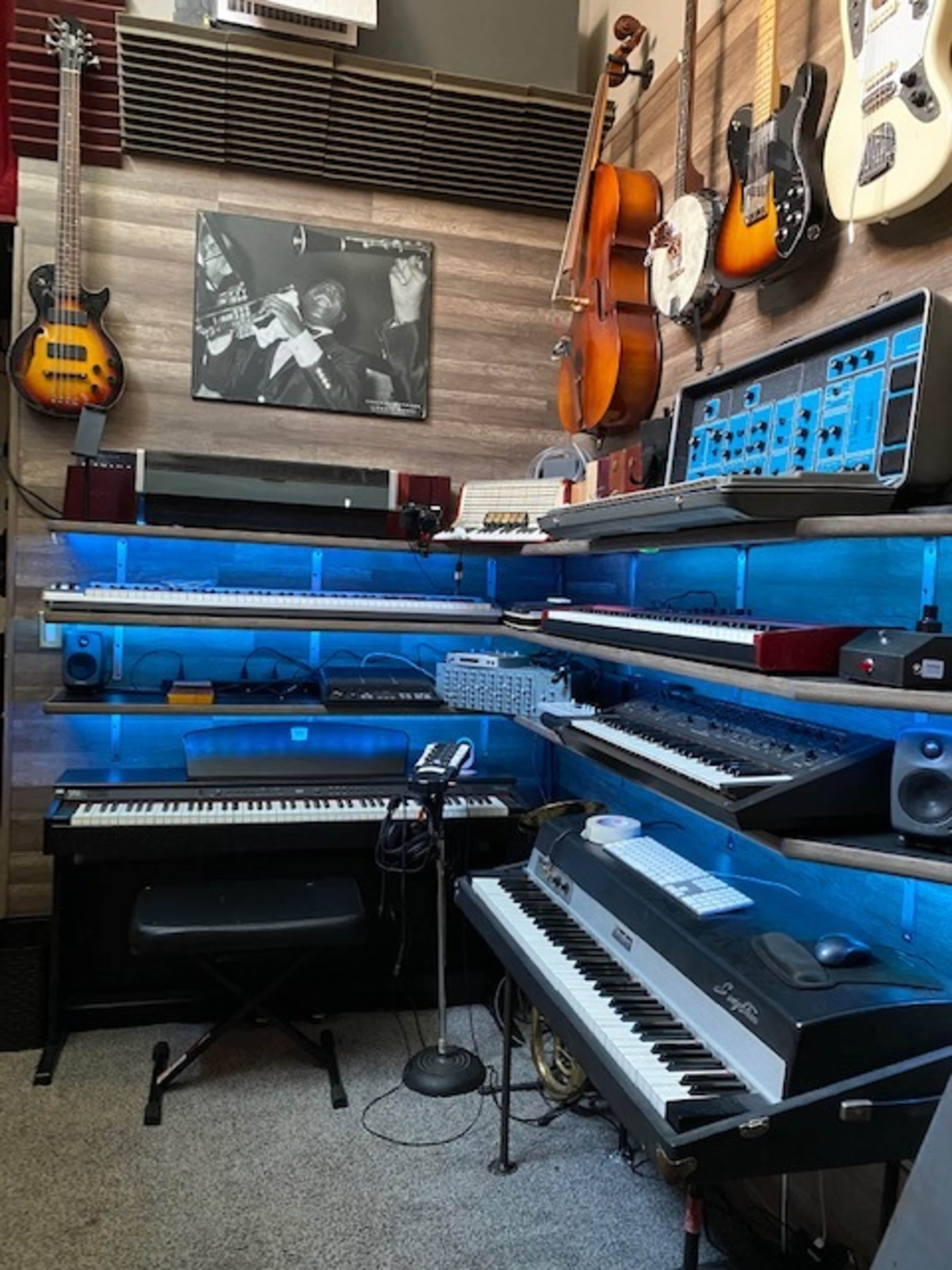 A music studio filled with various musical instruments, including keyboards, synthesizers, and guitars, arranged on multiple levels against a wooden wall.