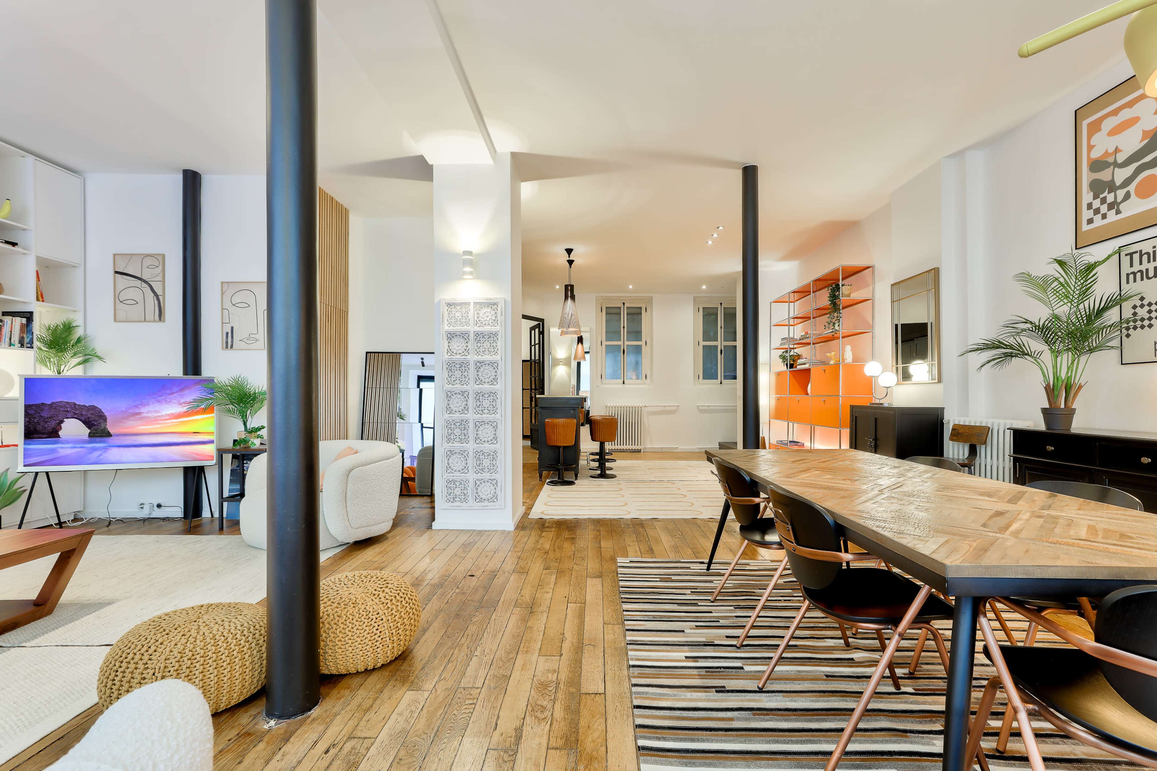 A spacious, modern living area with a wooden dining table, black chairs, a large television, and decorative shelving, featuring a mix of textures and colors.