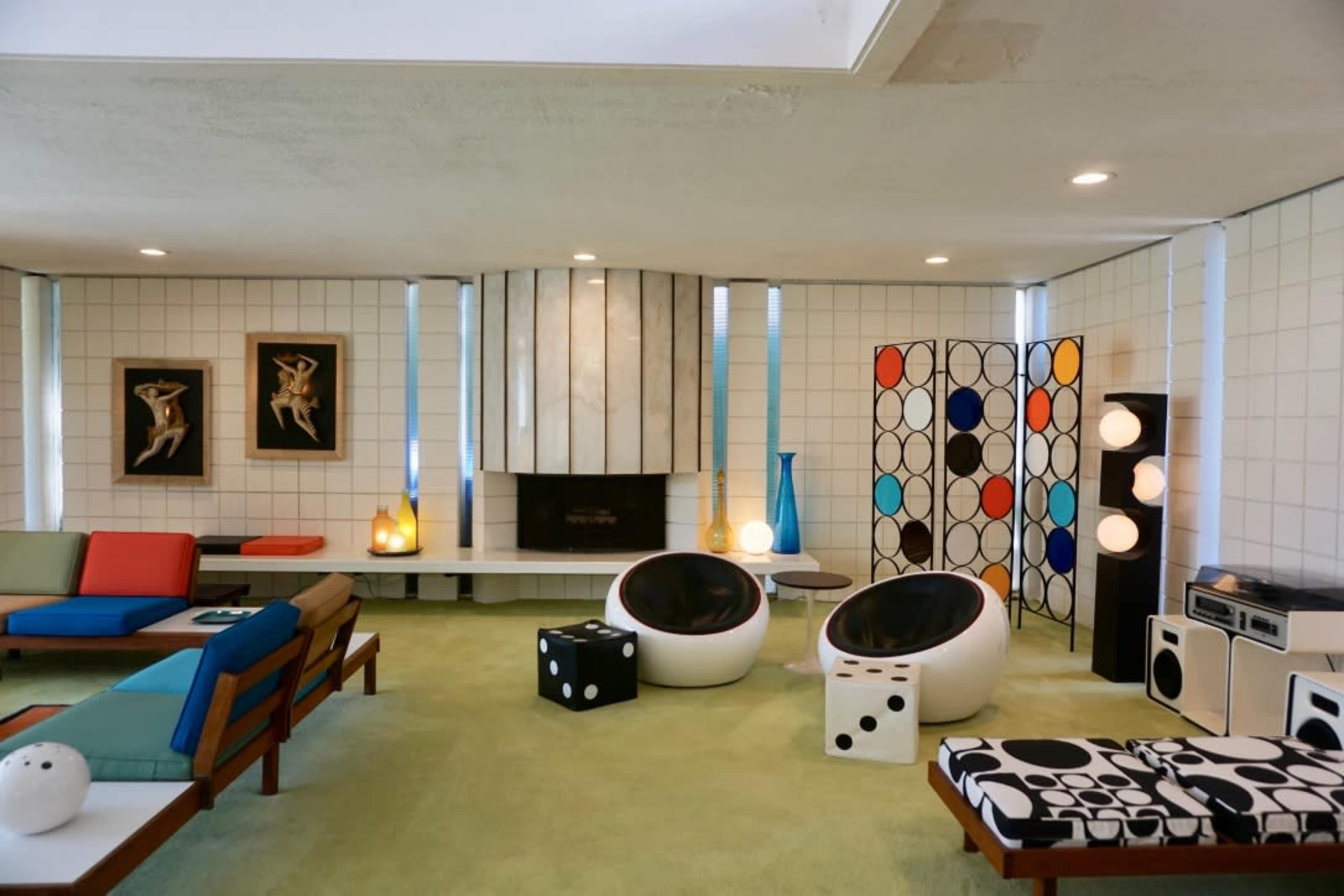 The room features a contemporary design with a green carpet, various seating options, a fireplace, and colorful decor elements.