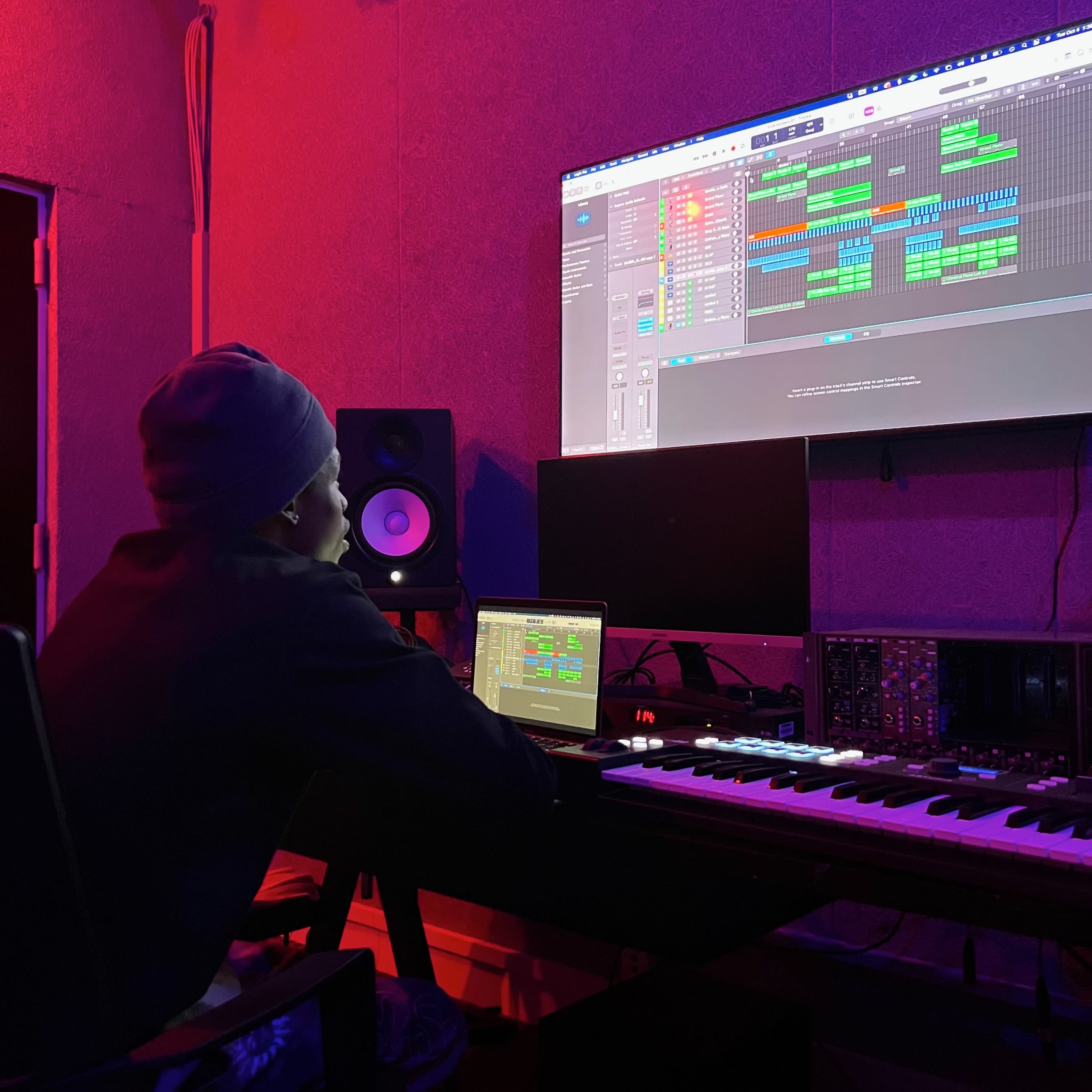 A person sits in a music studio, focused on a large screen displaying a digital audio workstation with a music project, surrounded by audio equipment and colorful lighting.