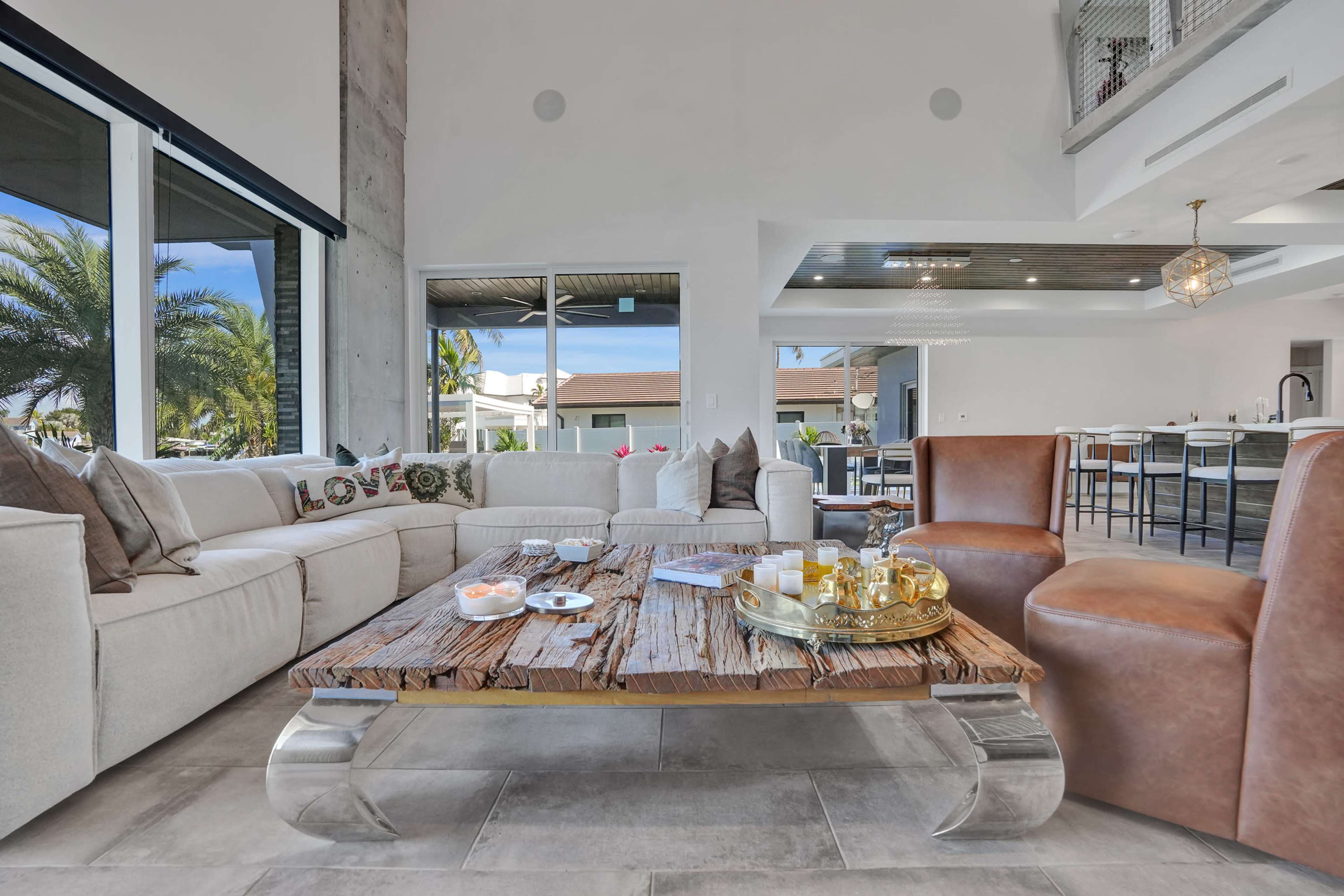 The image shows a modern living room featuring a beige sectional sofa, a wooden coffee table, and brown leather chairs, with large windows revealing a view of palm trees outside.
