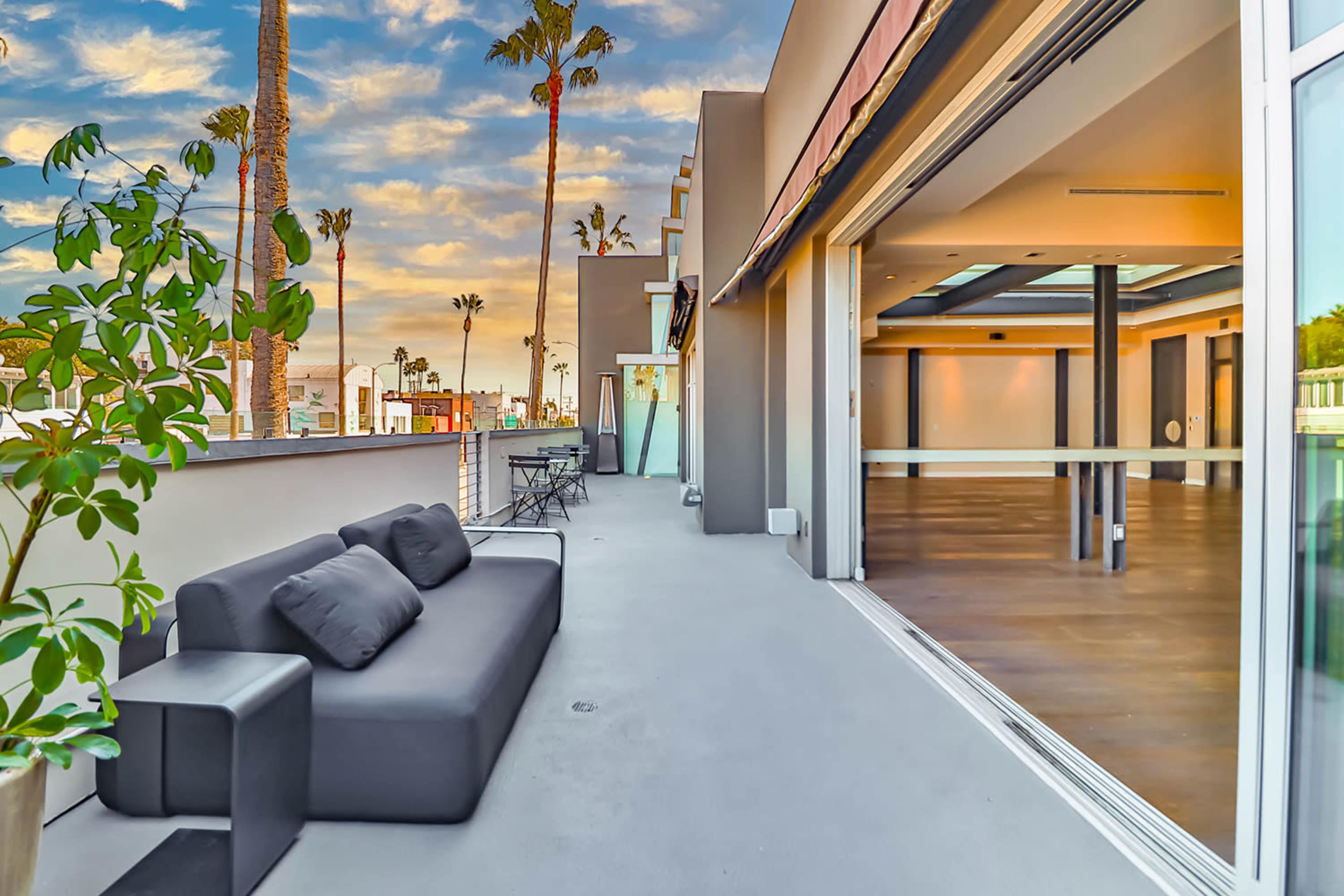 A spacious balcony features a gray sofa and overlooks palm trees against a colorful sky.