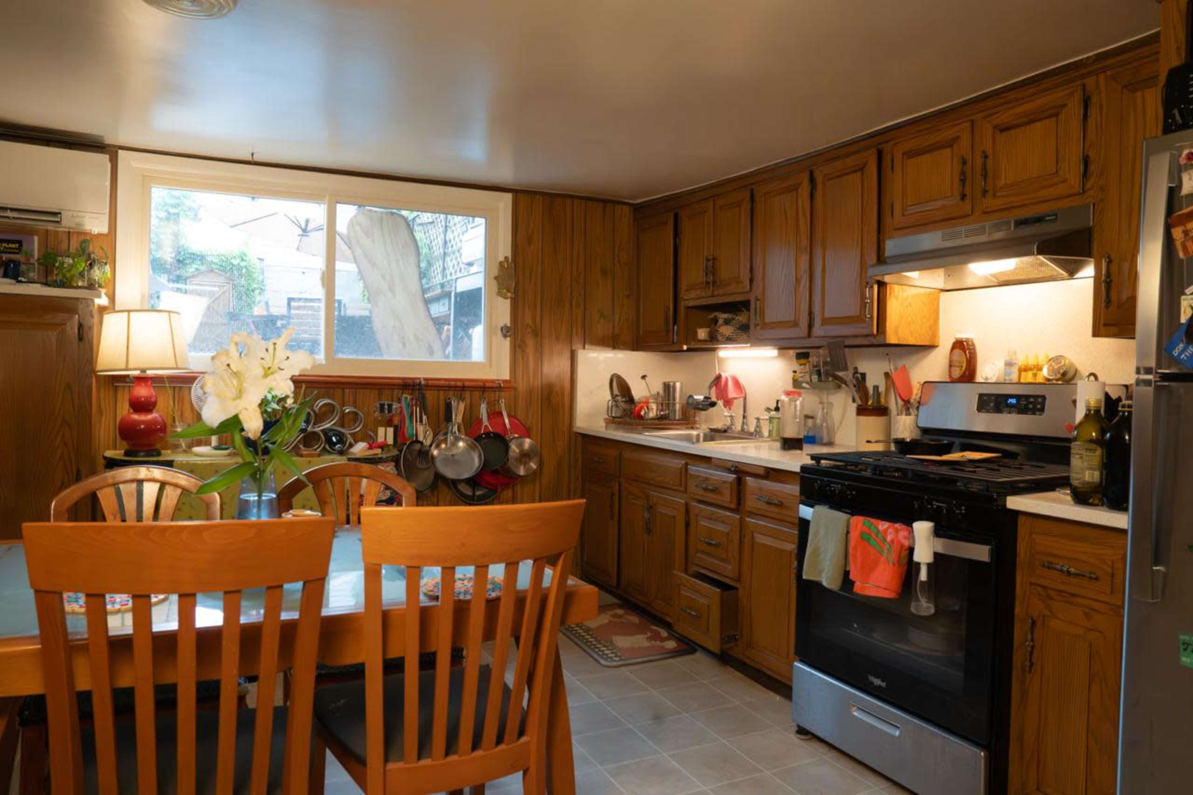 The image shows a cozy kitchen with wooden cabinets, a dining table set for four, and a view of a backyard through a large window.