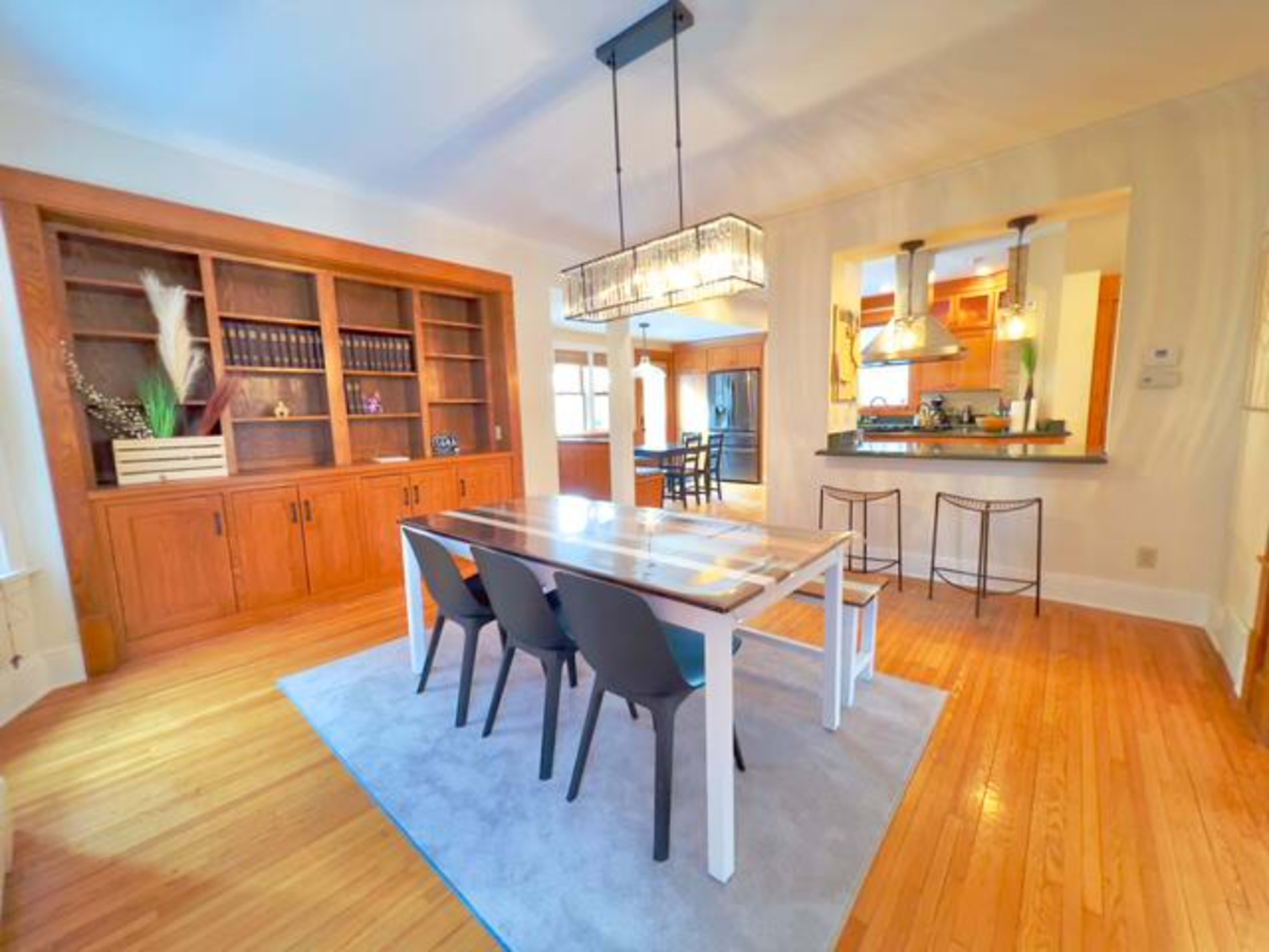 The image shows a modern dining area with a rectangular table surrounded by four chairs, a built-in wooden bookshelf, and a kitchen visible in the background.