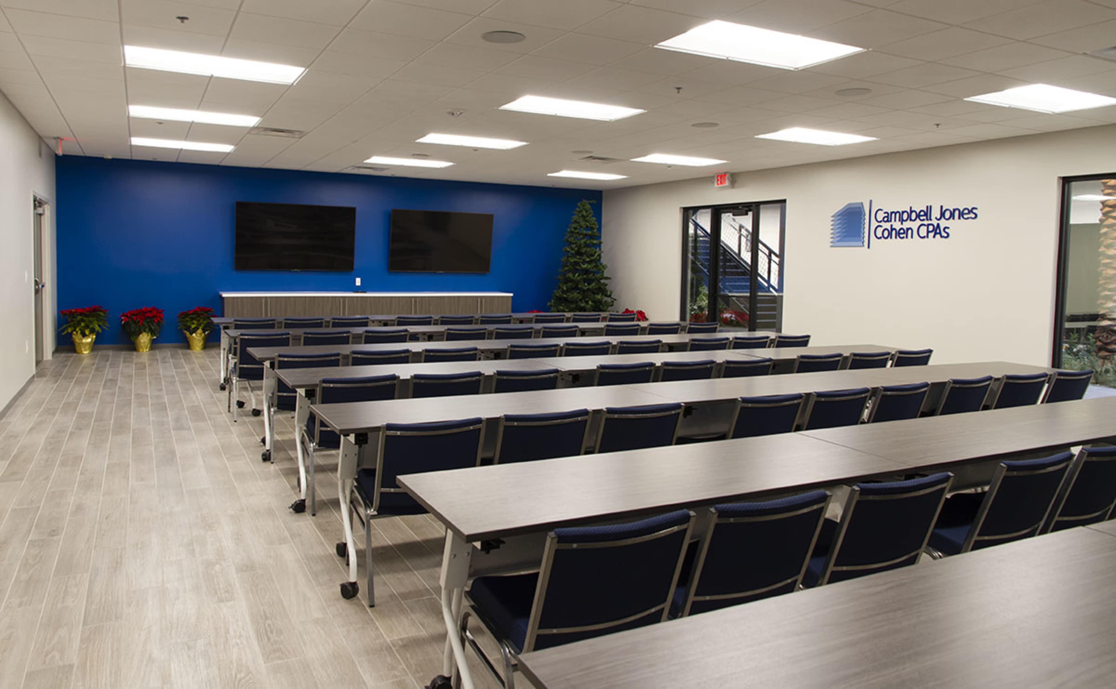 A modern conference room with rows of tables and chairs facing a wall featuring two screens and a company logo.