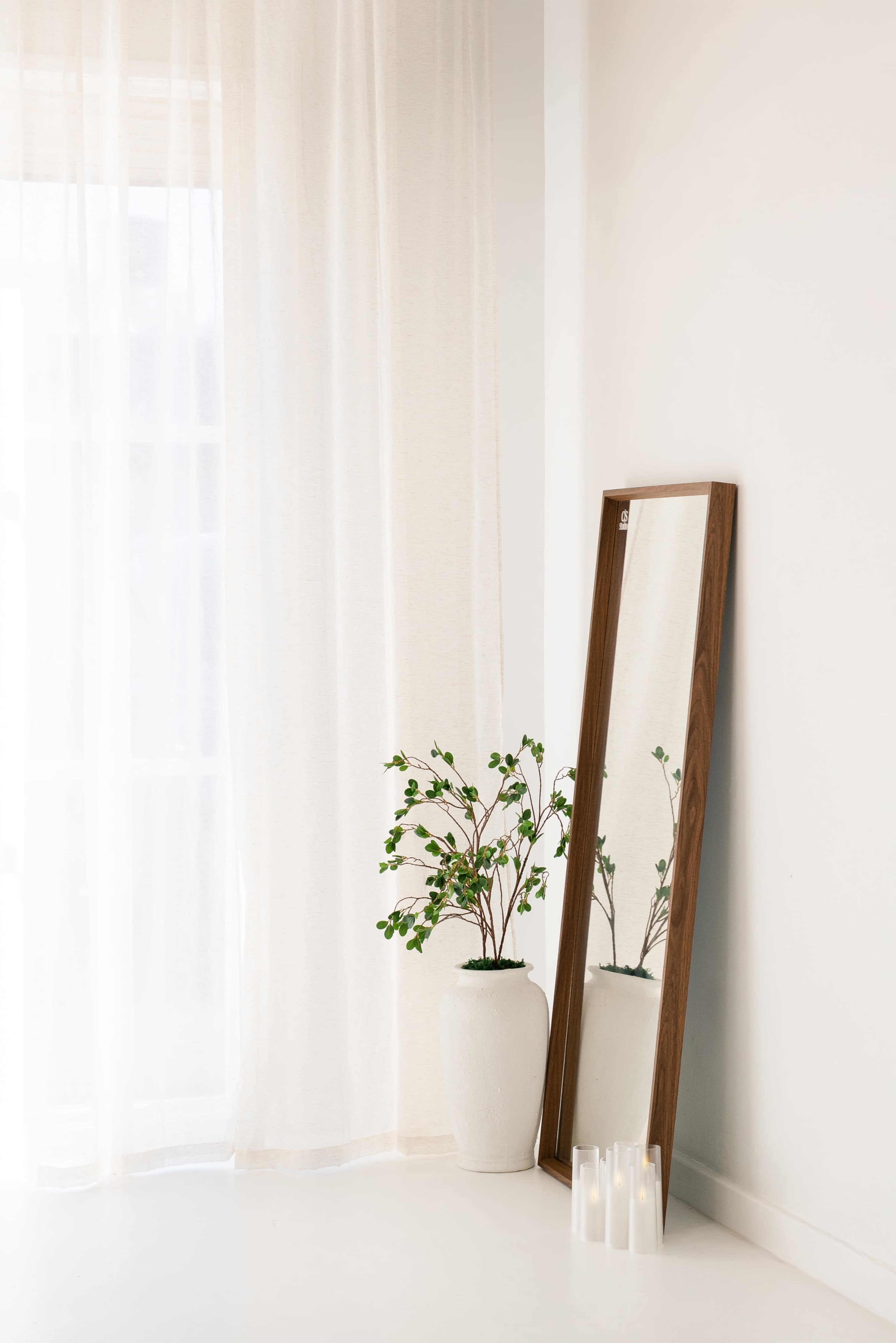 The image shows a tall mirror leaning against a white wall, with a potted plant nearby and sheer curtains allowing light to pass through.