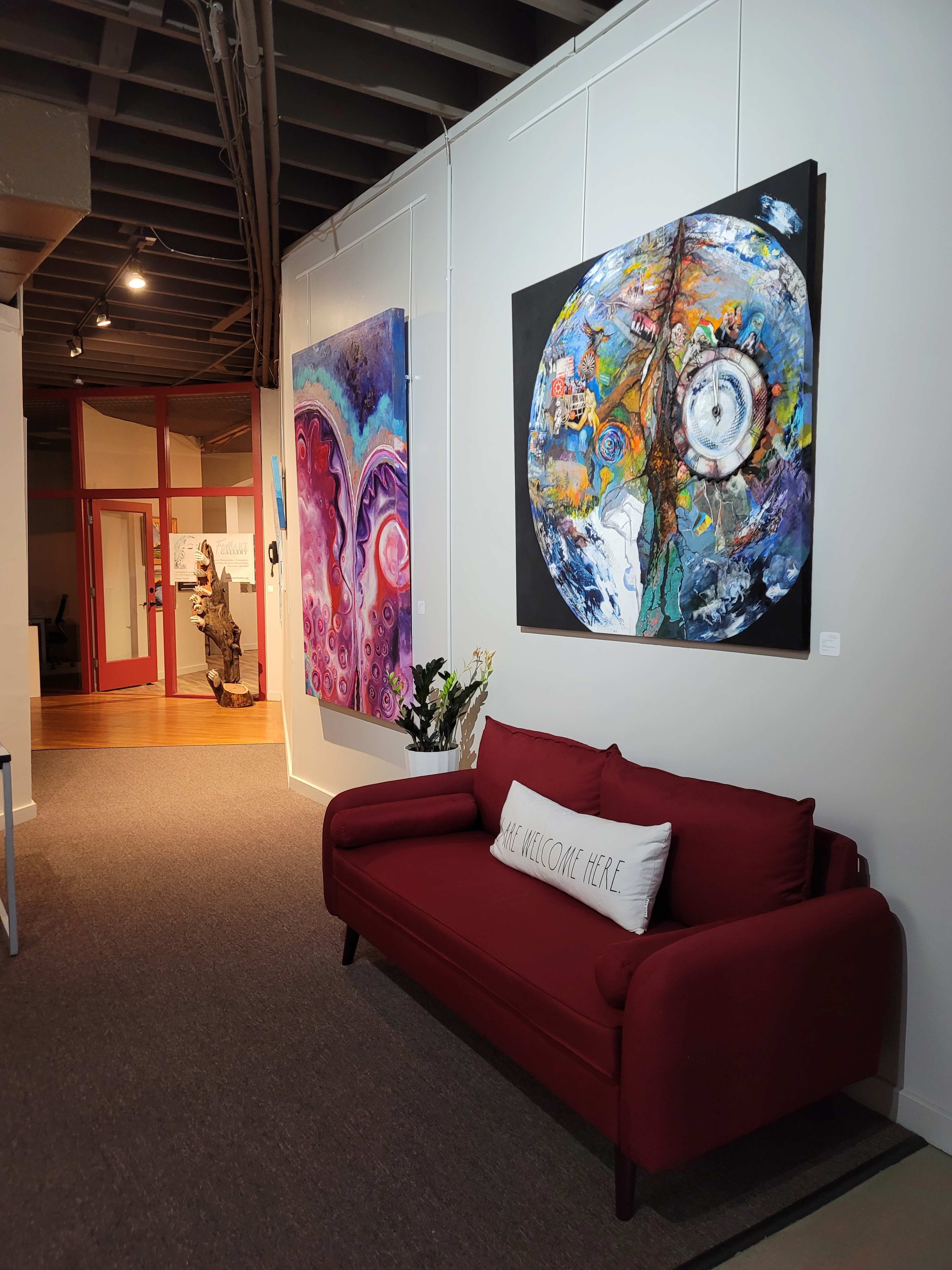 A gallery interior with colorful abstract paintings displayed on the walls and a red couch in the foreground, featuring a decorative pillow that reads "WE WELCOME HERE."