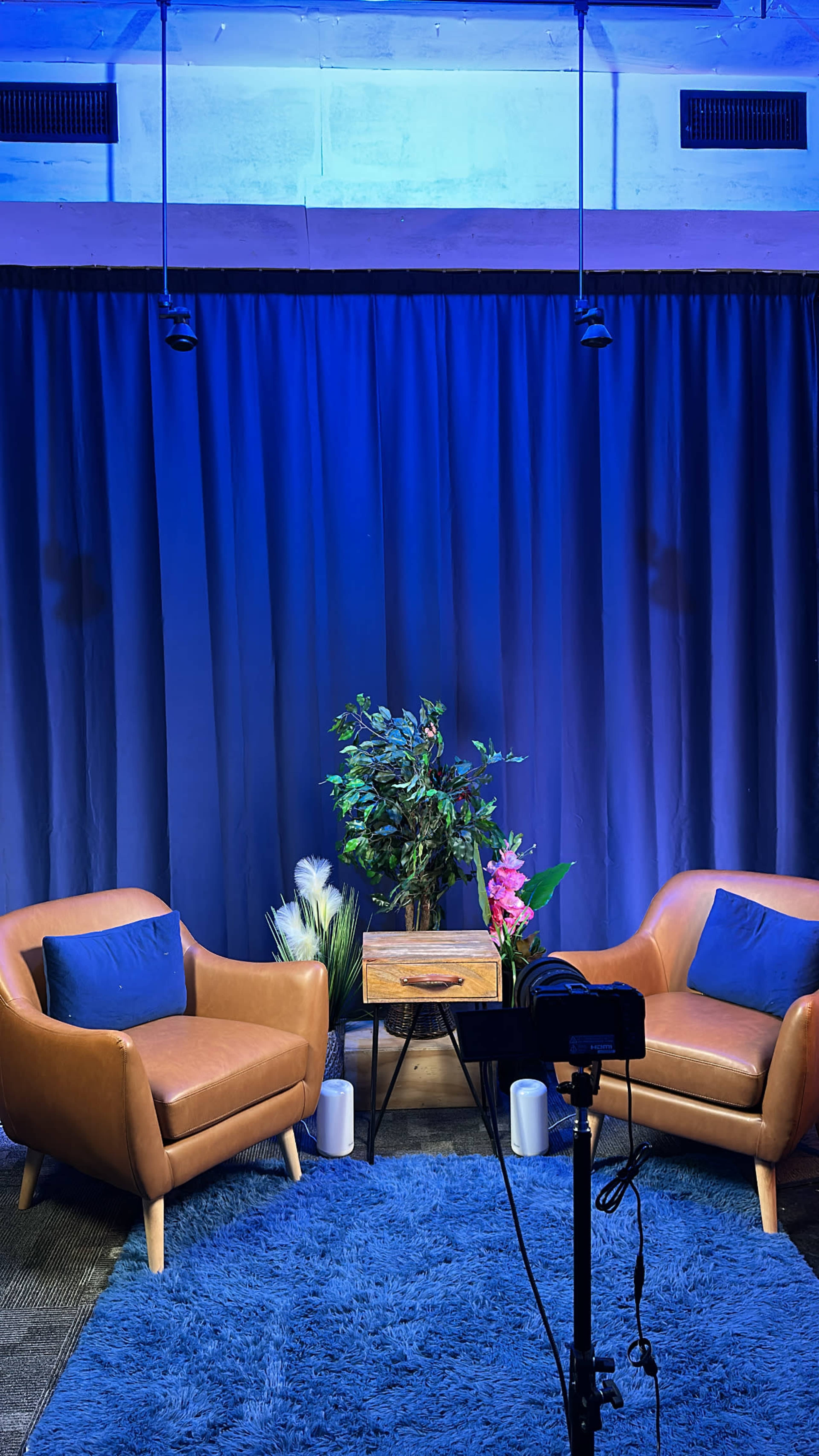 The image shows a modern interview setup with two brown chairs, a small wooden table, and decorative plants against a blue-lit backdrop.