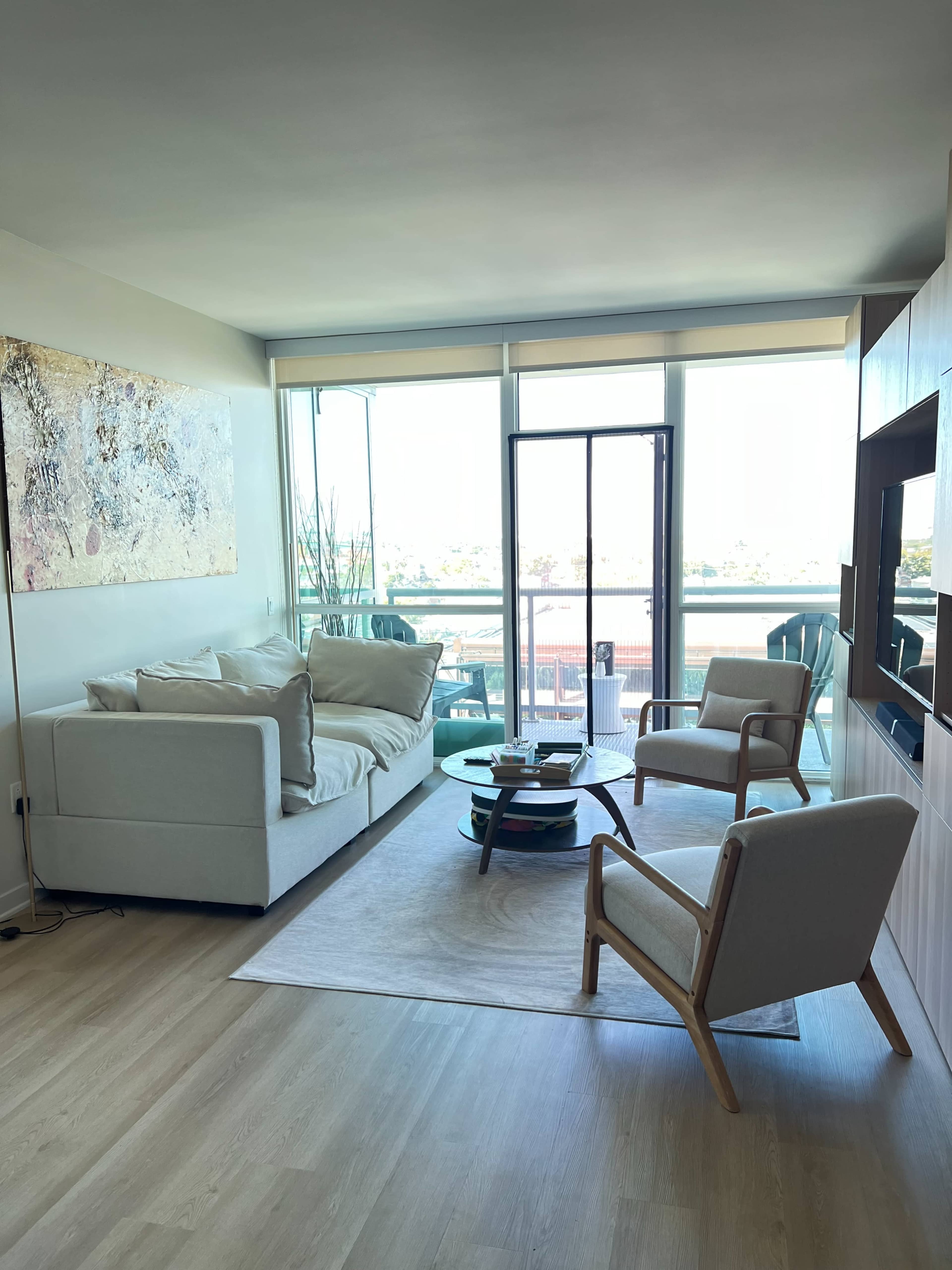 The image shows a modern living room with a white couch, two light-colored armchairs, a round coffee table, and large windows providing a view of the outdoors.