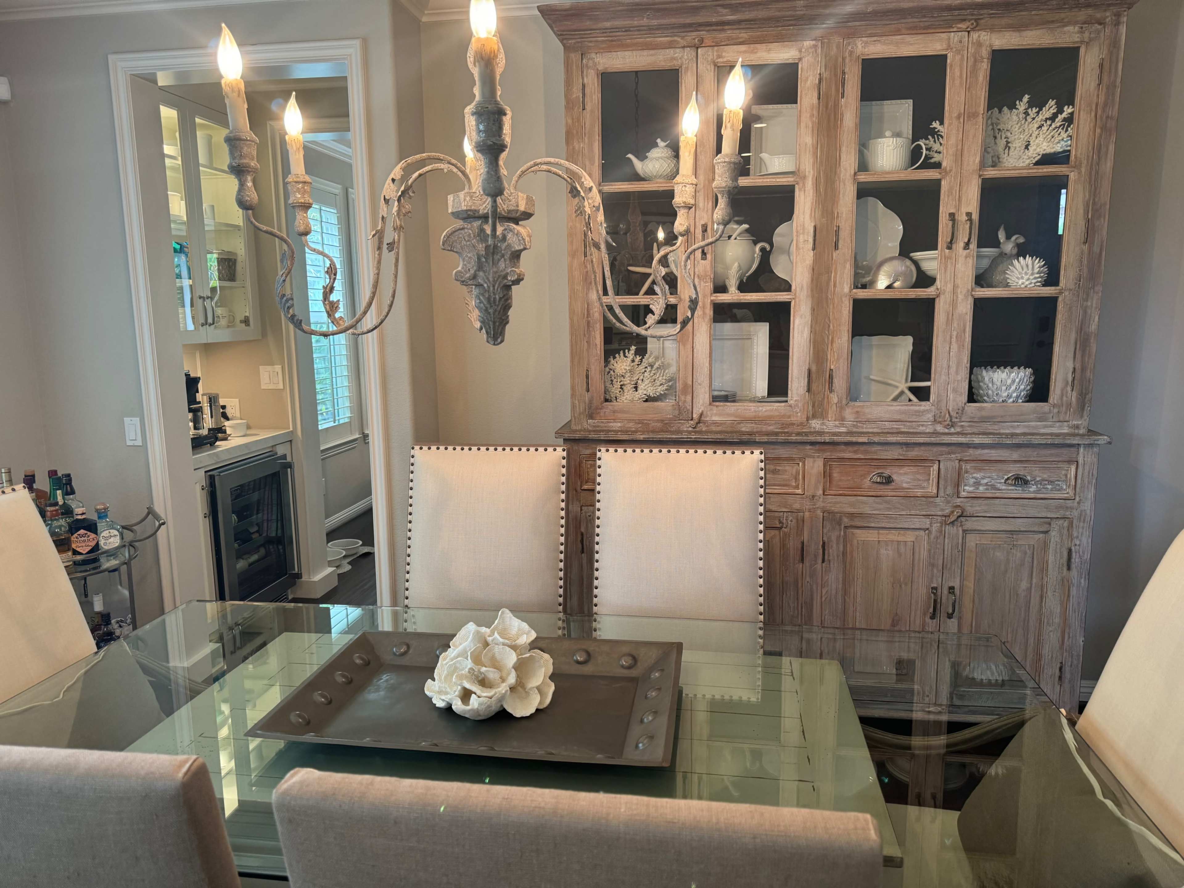 A dining area features a glass table with a central decorative item and a tall chandelier, complemented by beige upholstered chairs and a wooden cabinet displaying dishware.