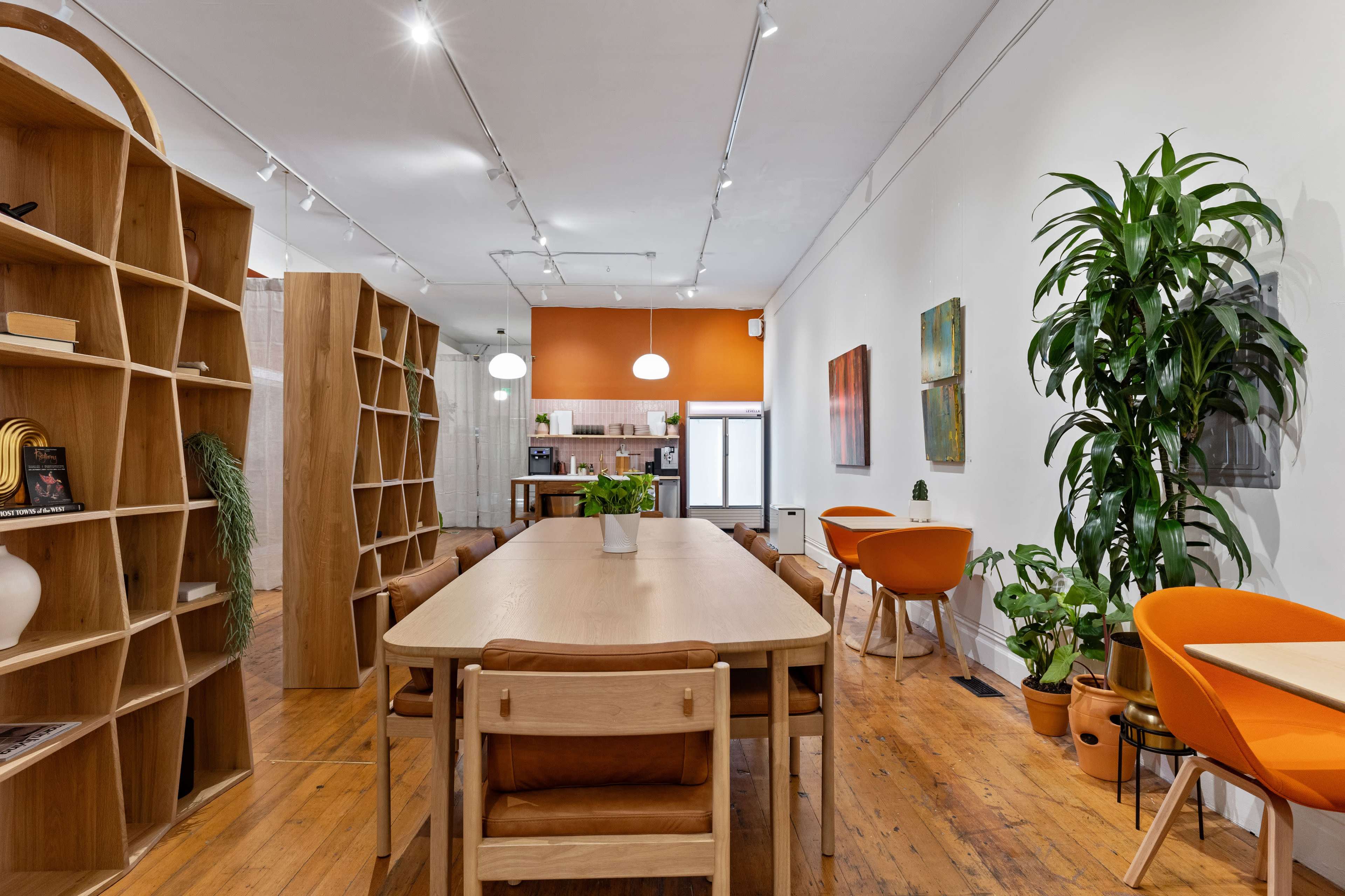 A modern dining area with a long wooden table, surrounded by orange chairs and bookshelves, and features a large potted plant in a well-lit space.