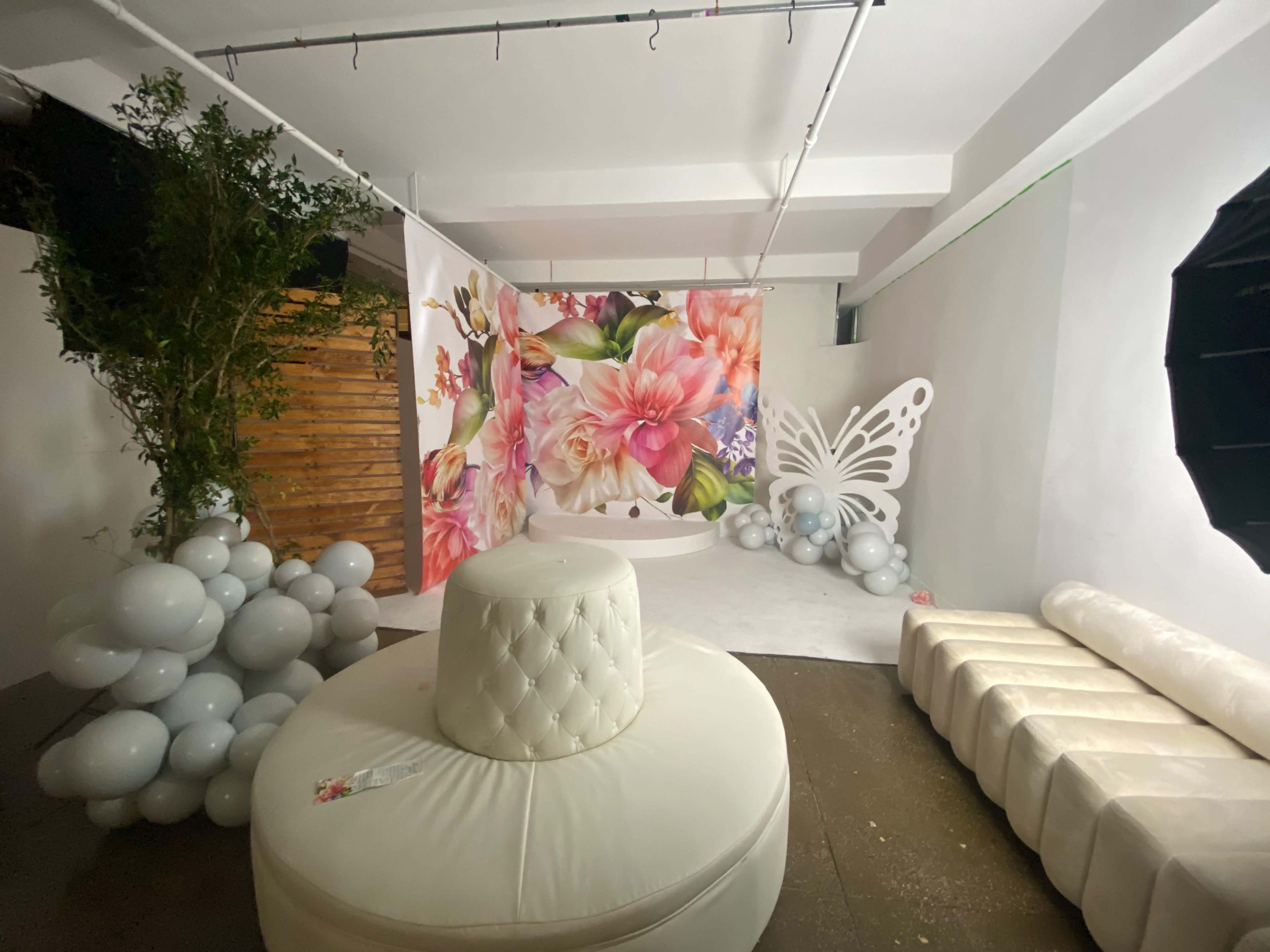 The image shows a decorated indoor event space featuring a floral backdrop, a large round white seating area, and clusters of balloons.