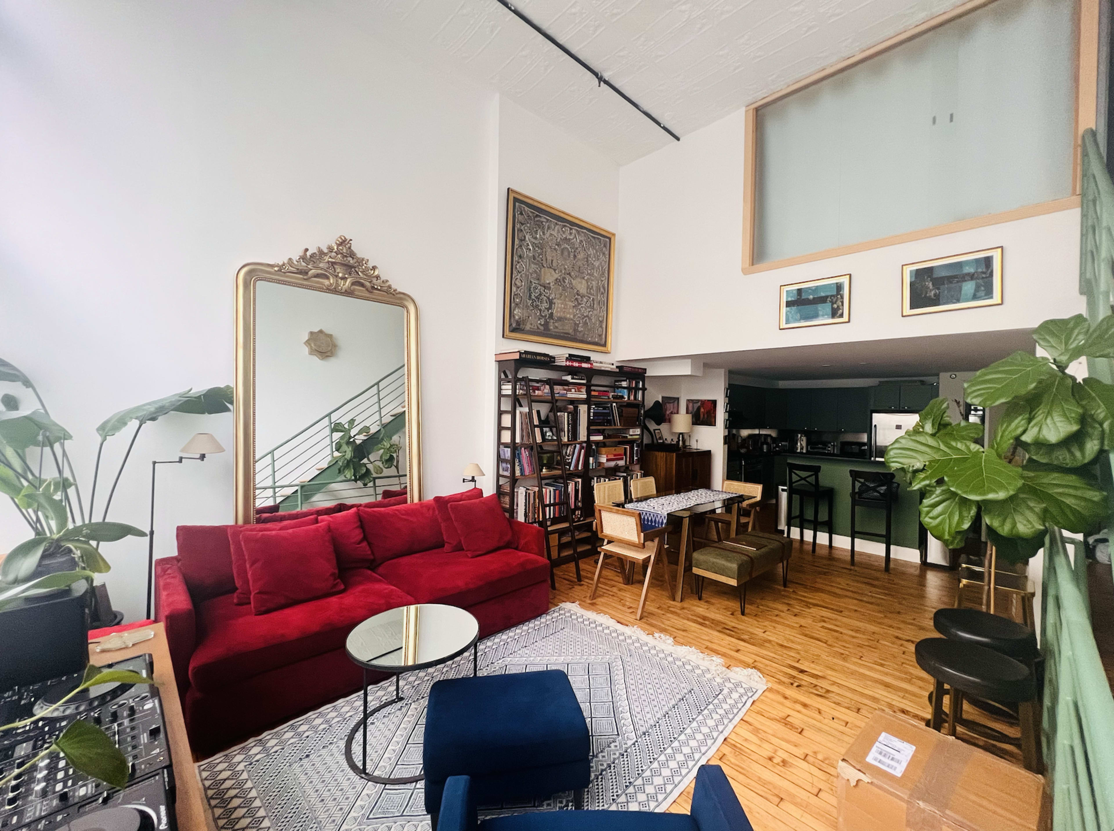 A bright, open living room featuring a red sectional sofa, a large mirror, a bookshelf filled with books, and a dining area with wooden furniture.