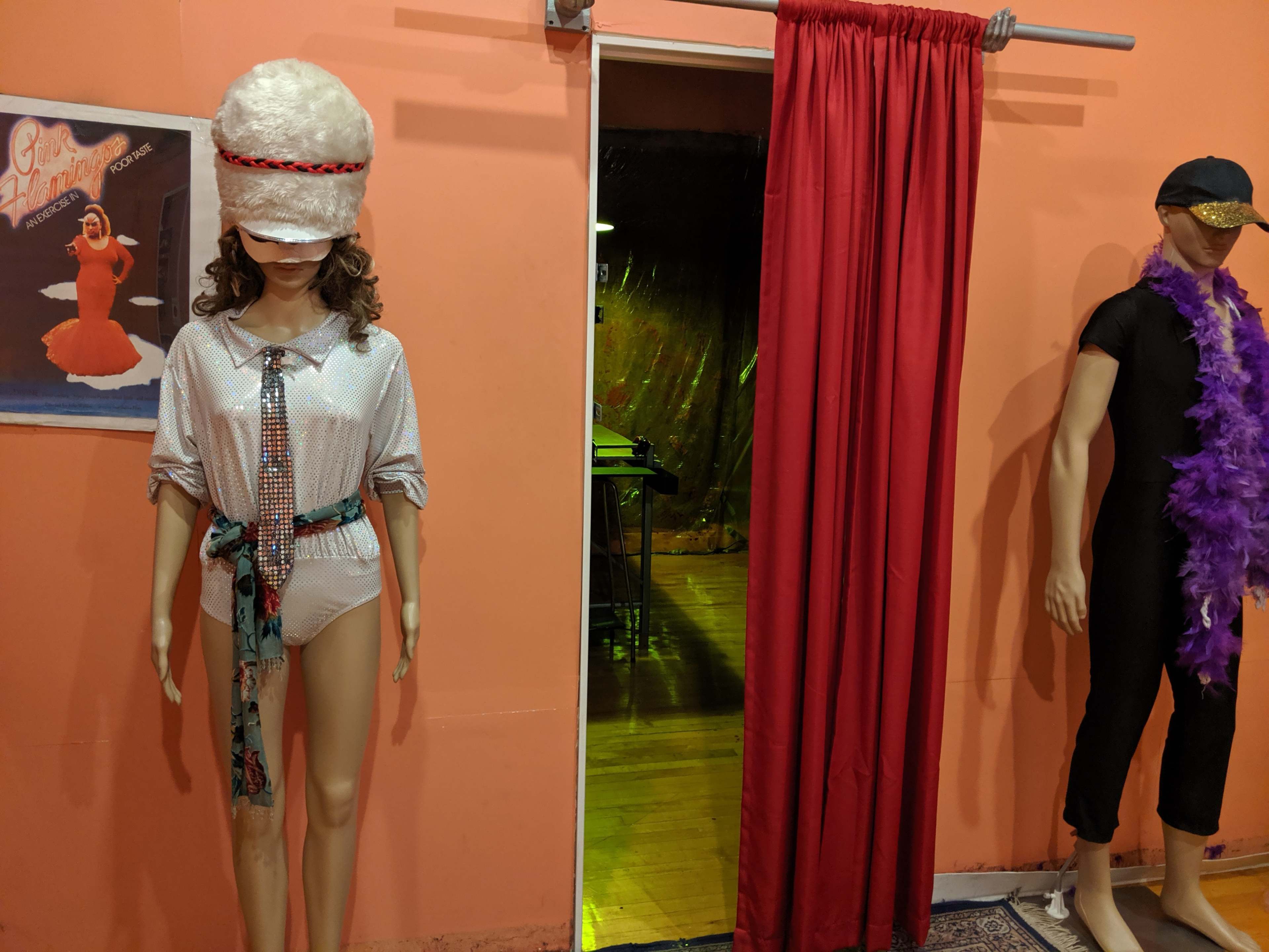 Two mannequins dressed in eccentric costumes, with one wearing a large fluffy hat and the other adorned with a feather boa, set against an orange wall and a red curtain.