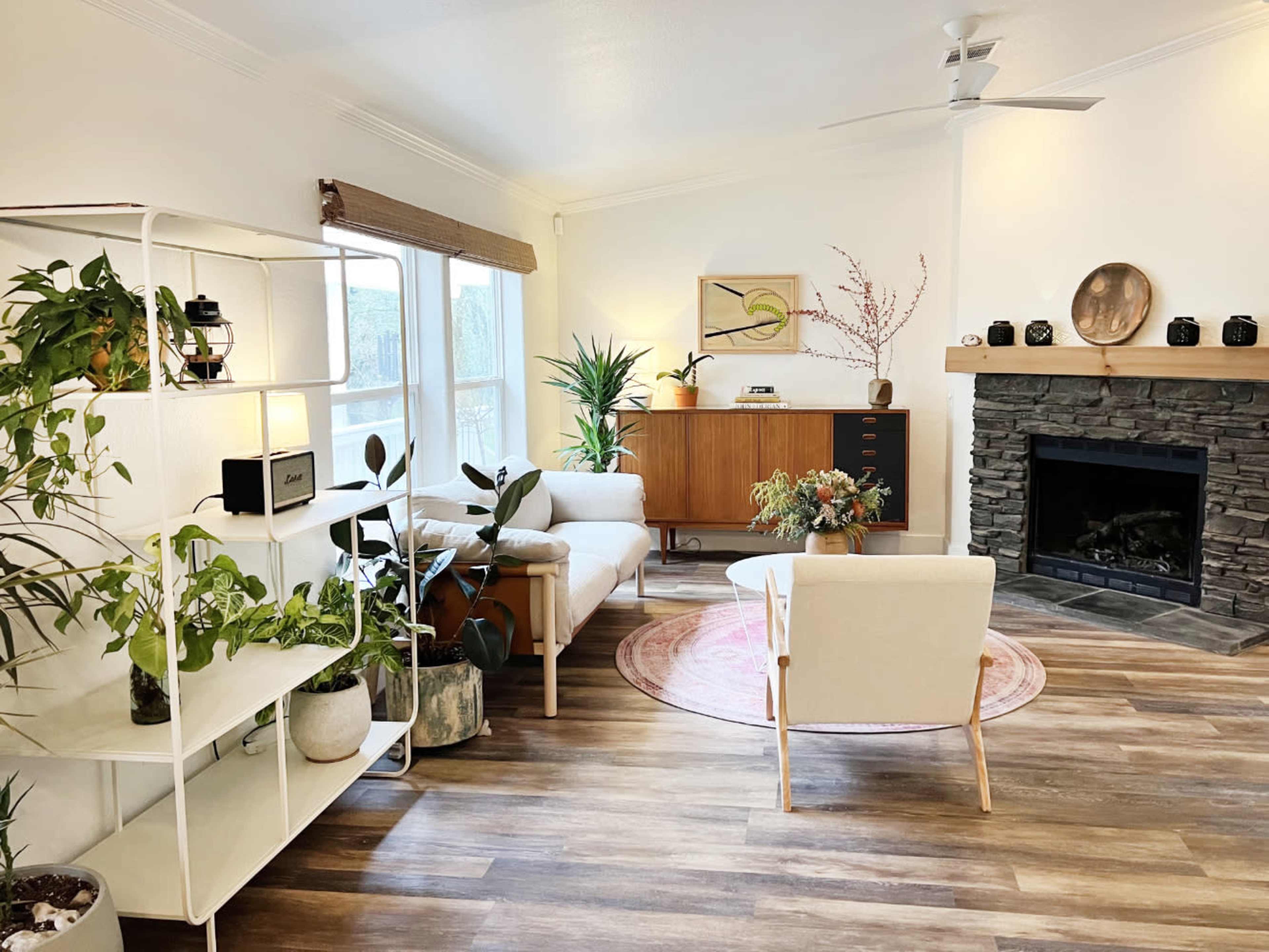 The image shows a modern living room with a white sectional sofa, a wooden sideboard, large windows, a stone fireplace, and various indoor plants arranged around the space.