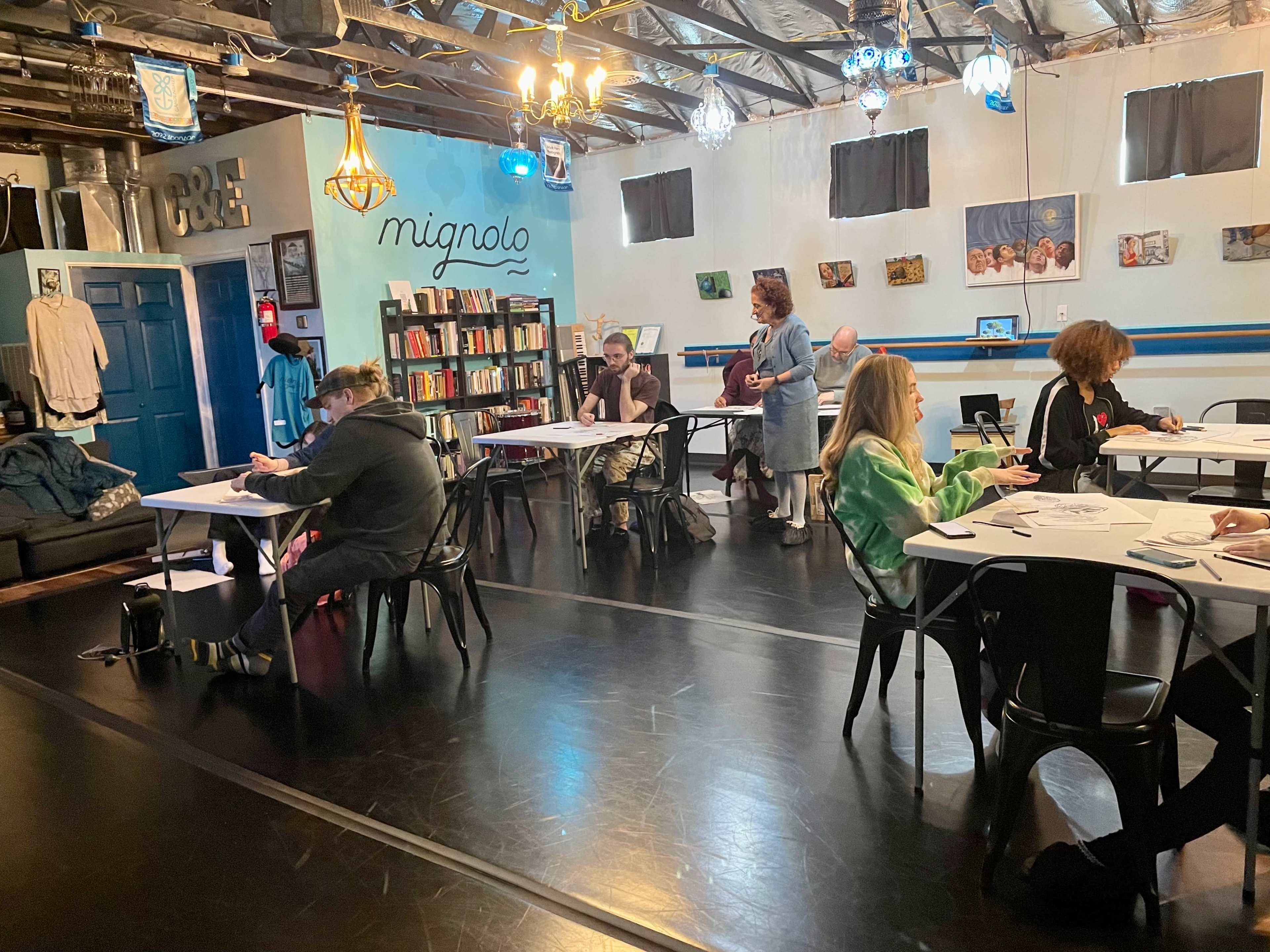 A group of people are seated at tables in a brightly lit room with bookshelves and artworks on the walls, engaged in various activities.