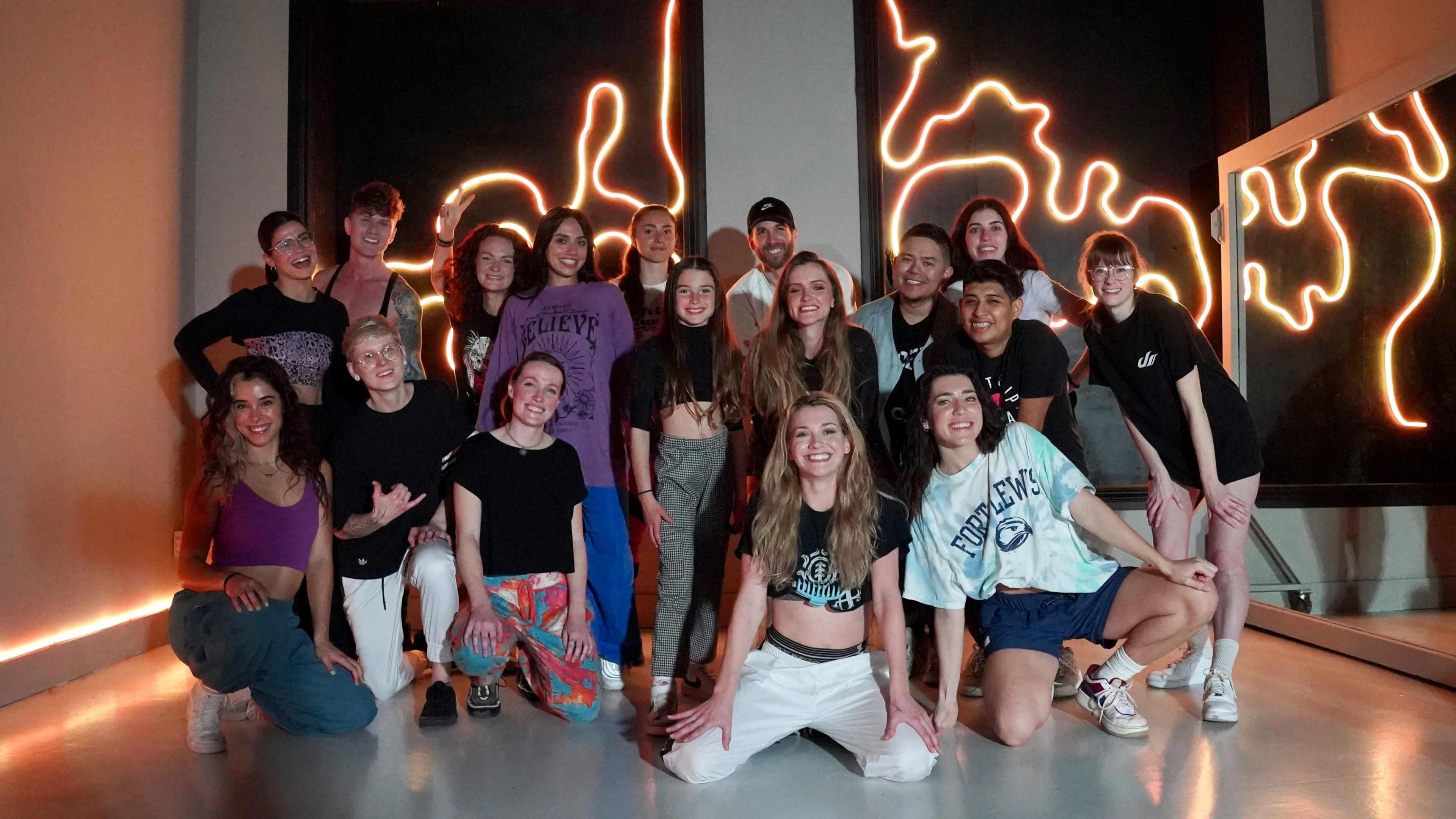 A group of twenty individuals poses together in a dance studio with neon lighting in the background.