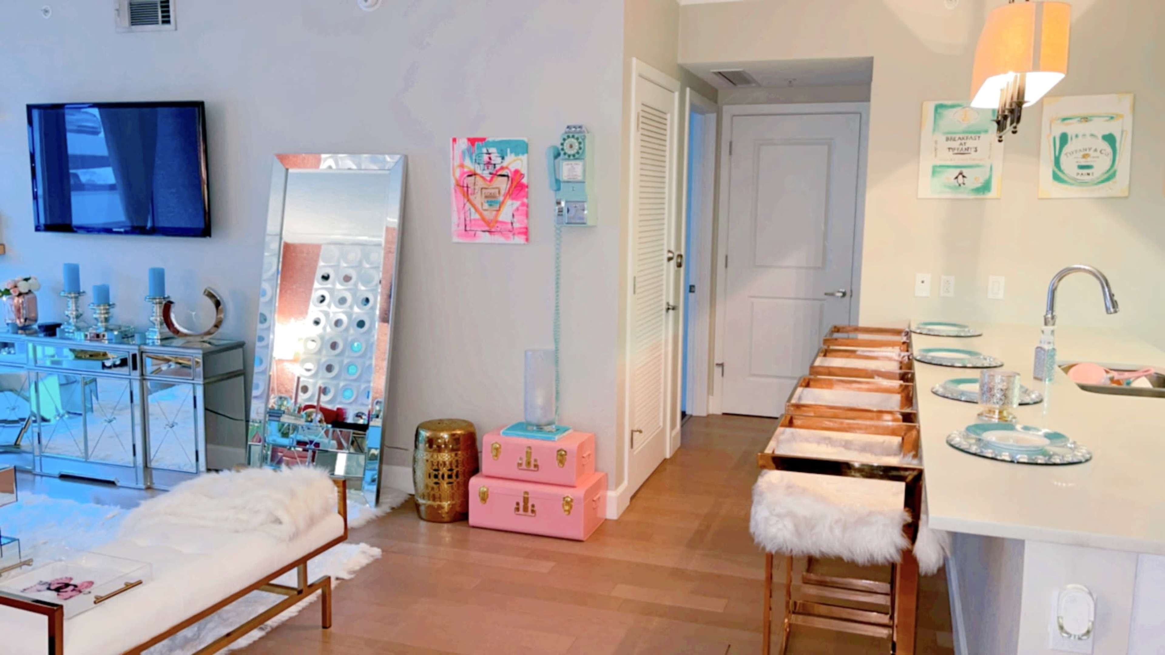 A bright, modern living space featuring a large mirror, a pink storage unit, and a kitchen bar with wooden stools.