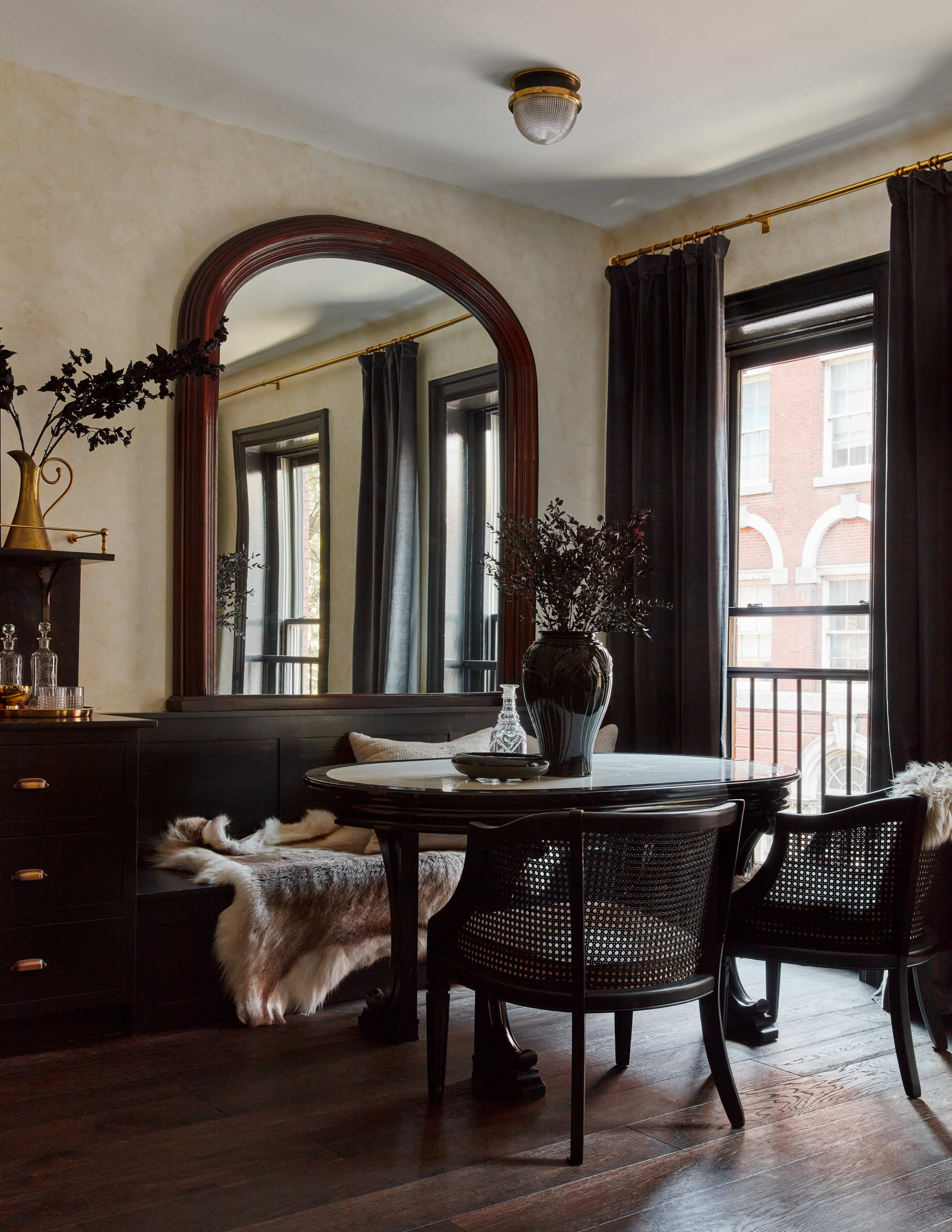 A dining area features a round table with two chairs, a large mirror on the wall, and dark curtains framing the windows.