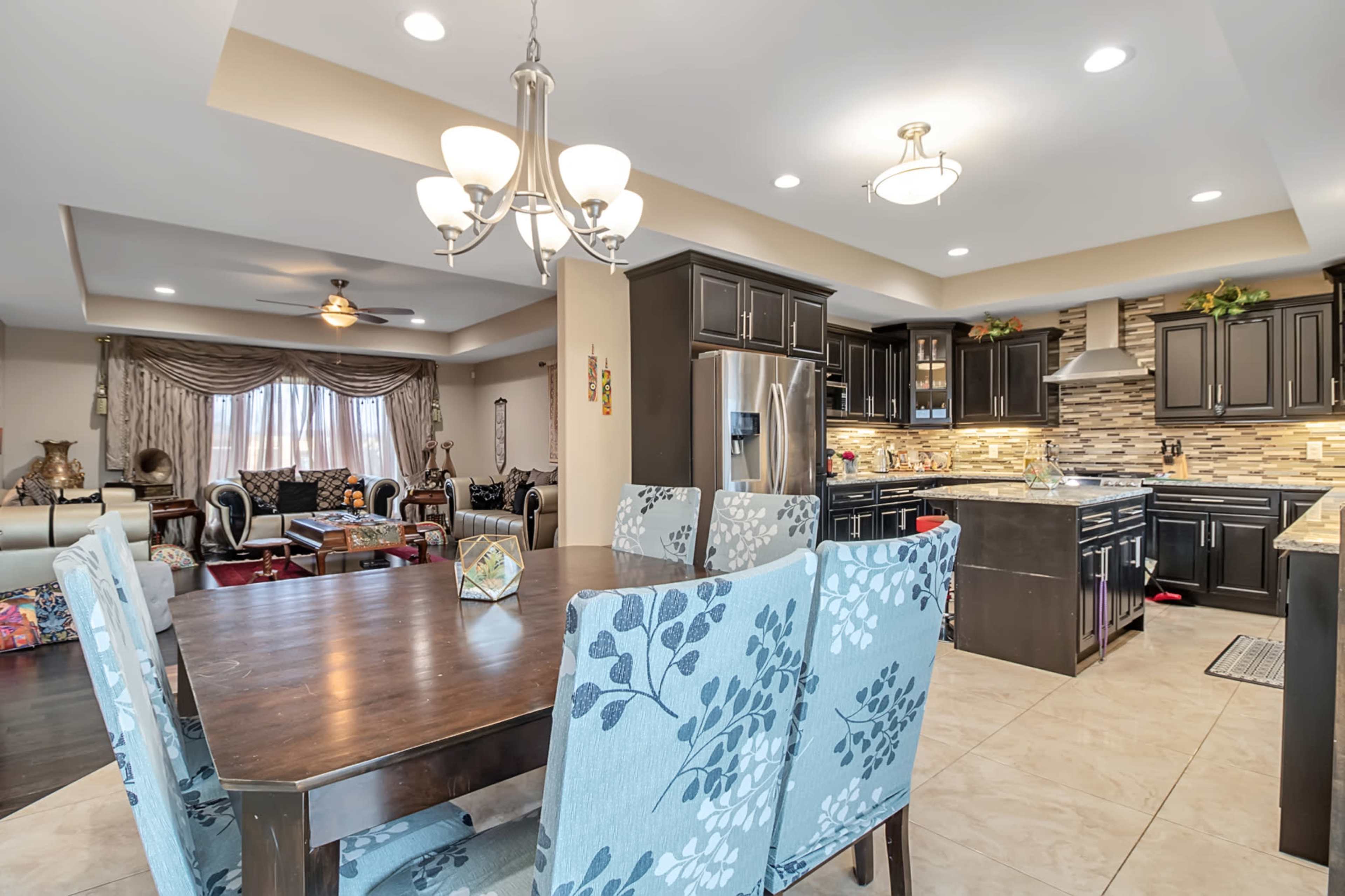 A modern kitchen and dining area with dark cabinetry, a large dining table with light blue chairs, and a living room visible in the background.