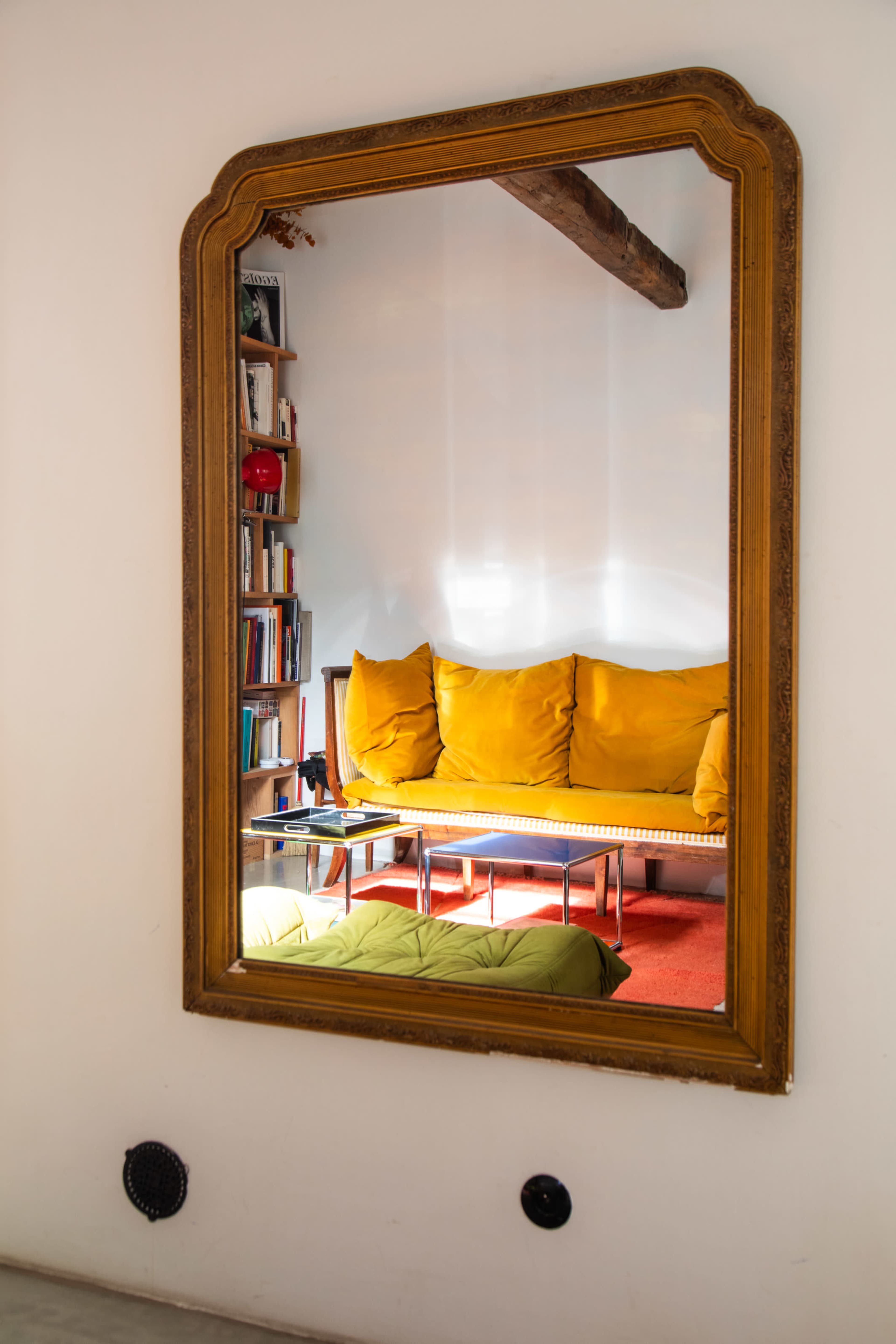 A large ornate mirror reflects a yellow sofa and a coffee table in a room with a bookshelf.