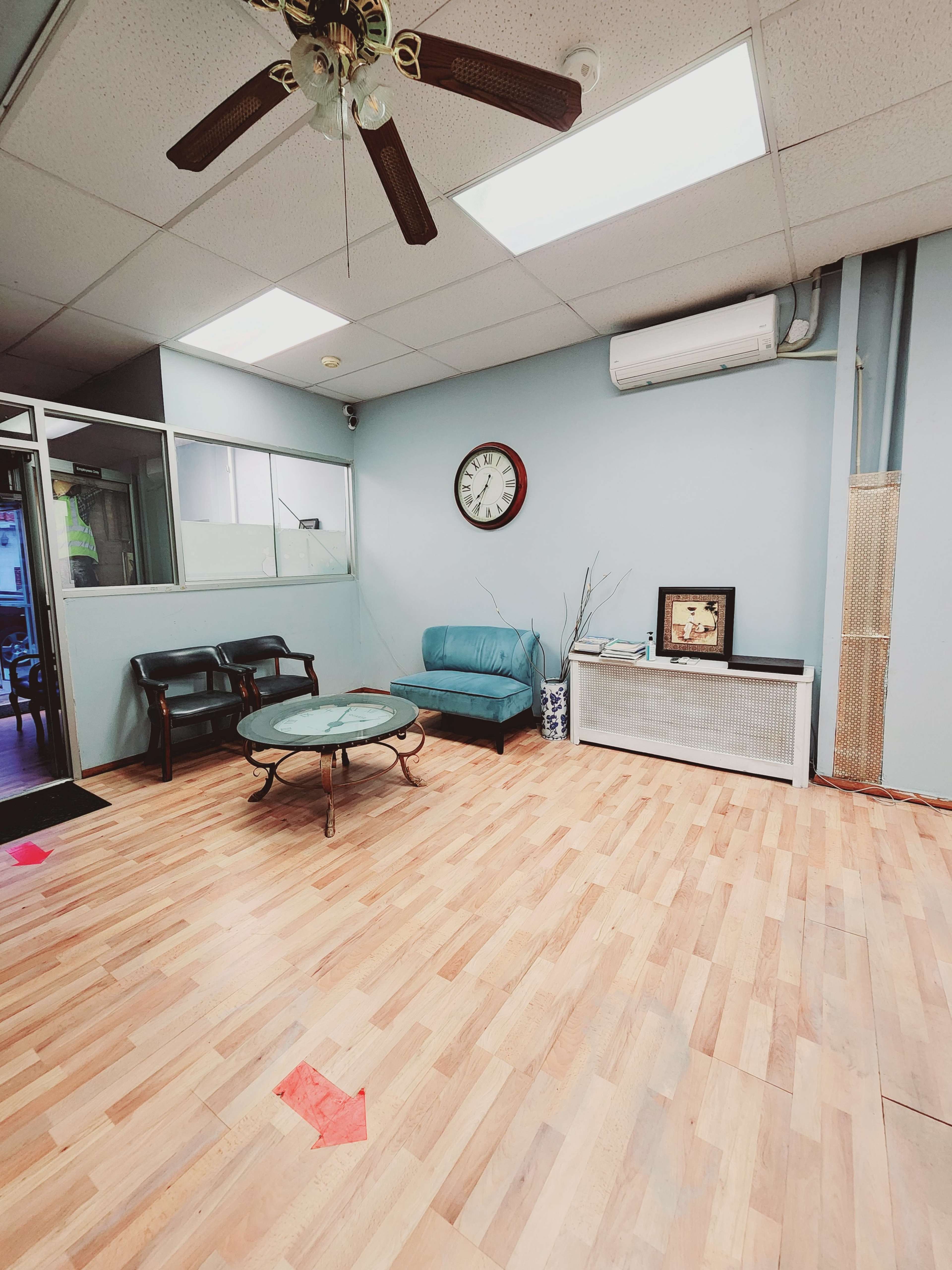 A waiting room with a blue sofa, a round glass coffee table, and a wall clock on a light blue wall, featuring a wooden floor and a visible air conditioning unit.