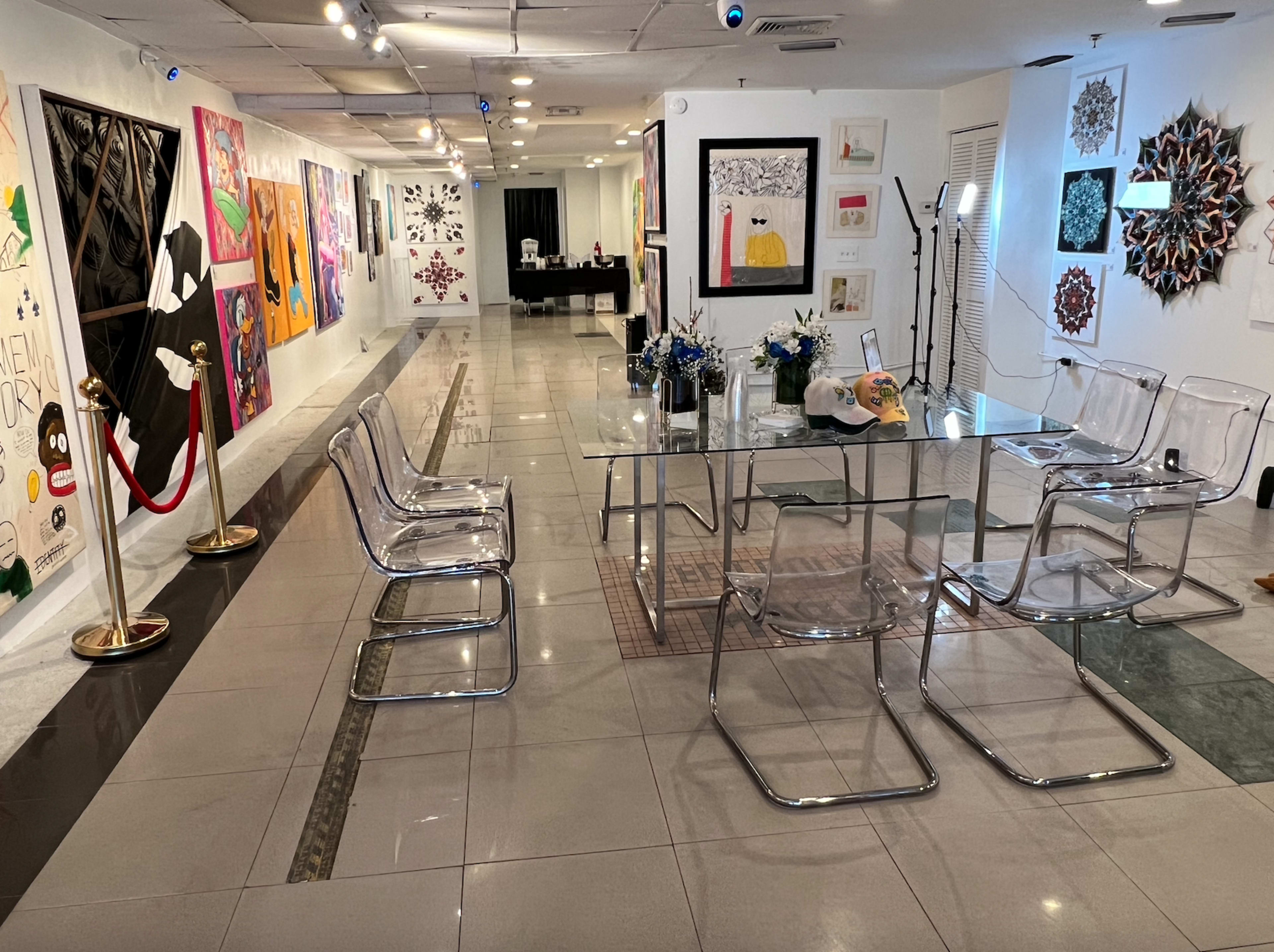A modern art gallery featuring a clear glass table surrounded by acrylic chairs, with colorful artworks displayed on the walls and a reception area in the background.