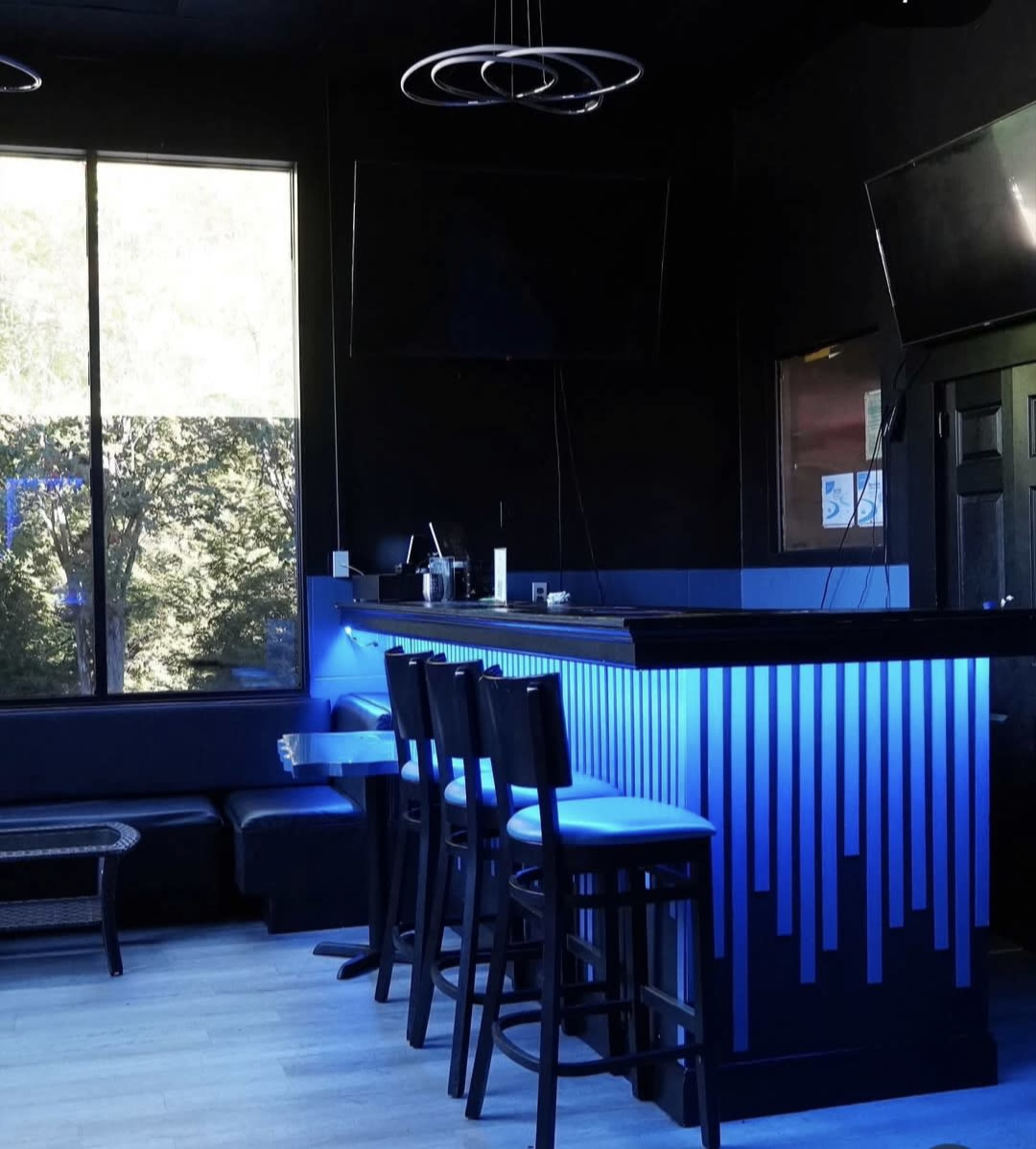The image shows a modern bar area with sleek black furnishings, illuminated by blue lighting, and several bar stools positioned in front of it.