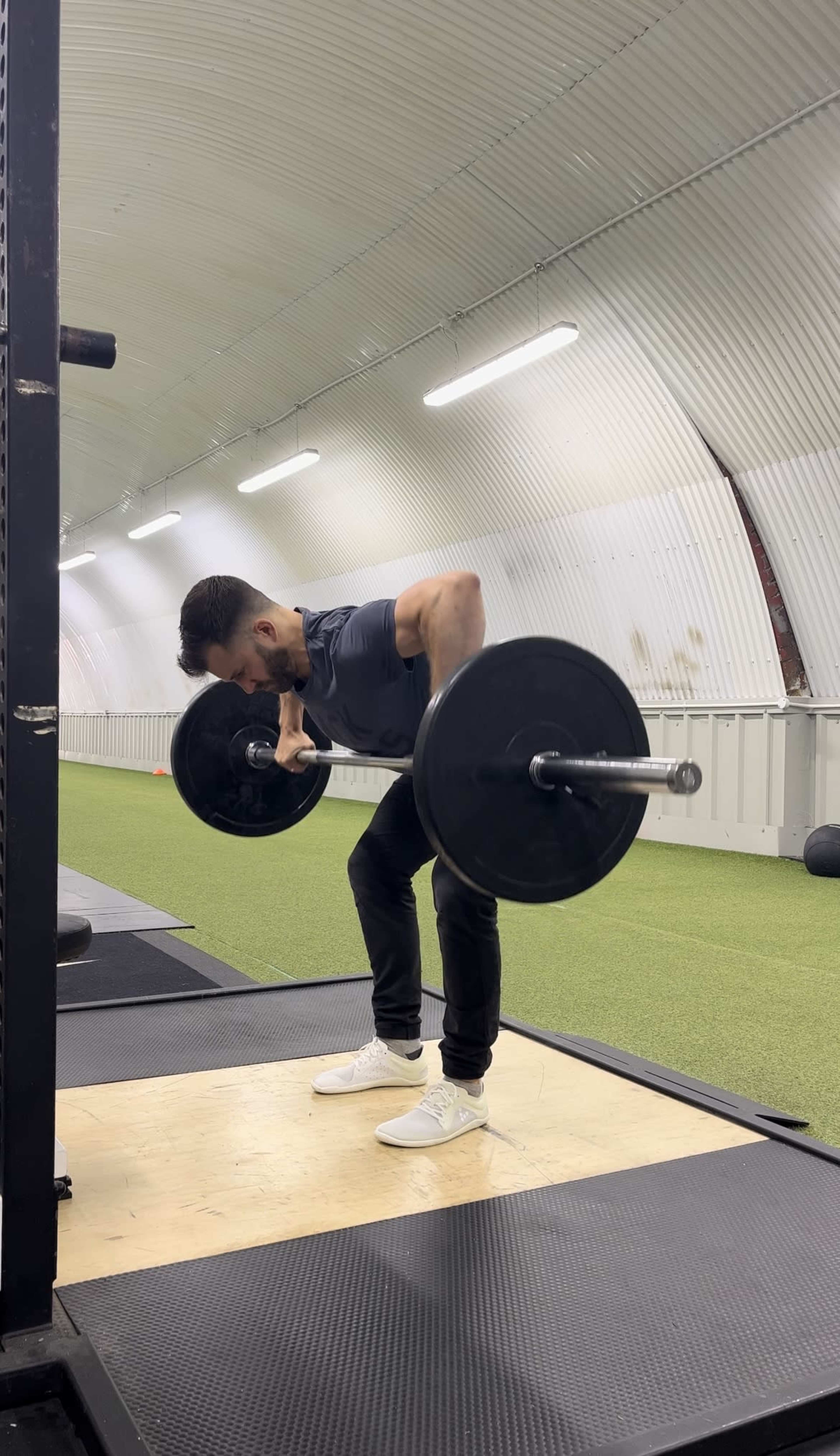 A person is performing a bent-over row with a barbell in a gym with a synthetic grass floor.