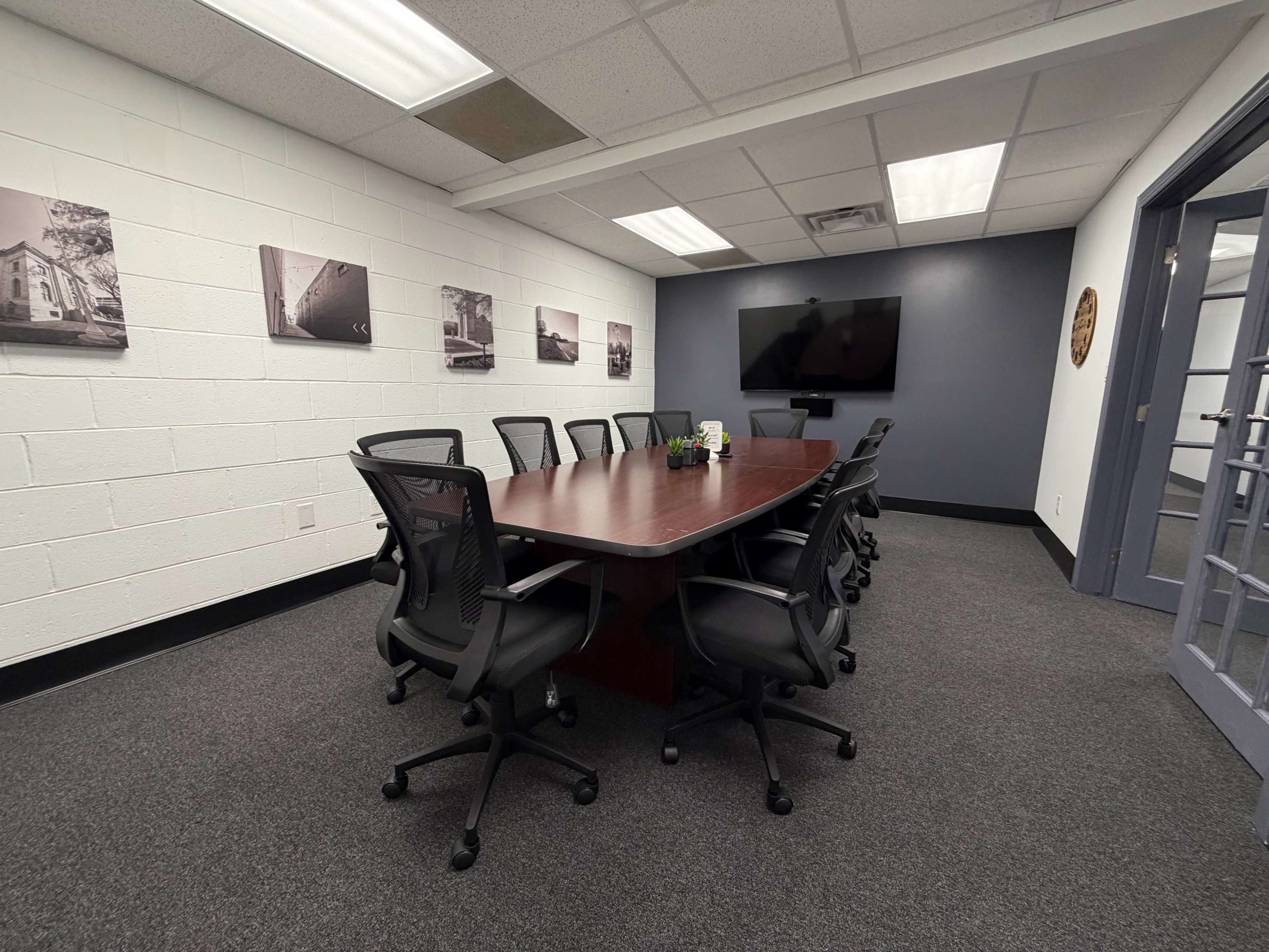The image shows a conference room with a large wooden table surrounded by black office chairs, and a wall featuring framed black-and-white photographs.