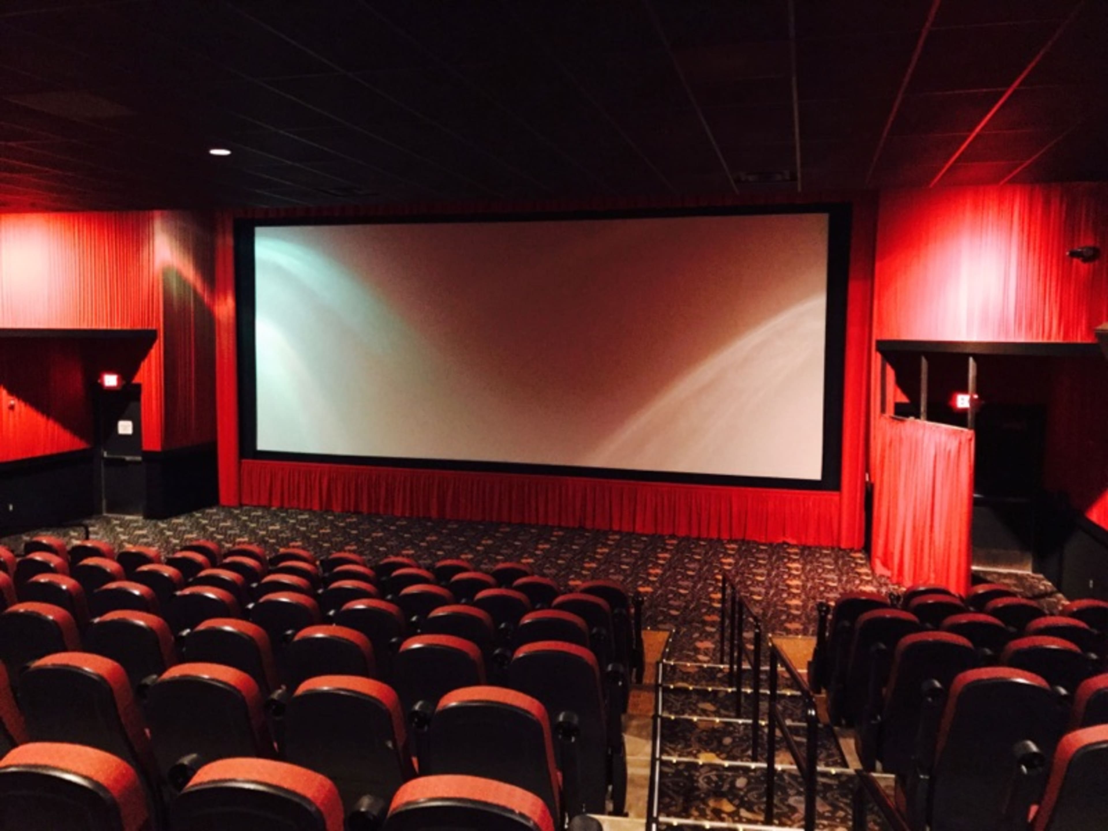 A nearly empty movie theater with red walls, a large screen, and rows of black seating.