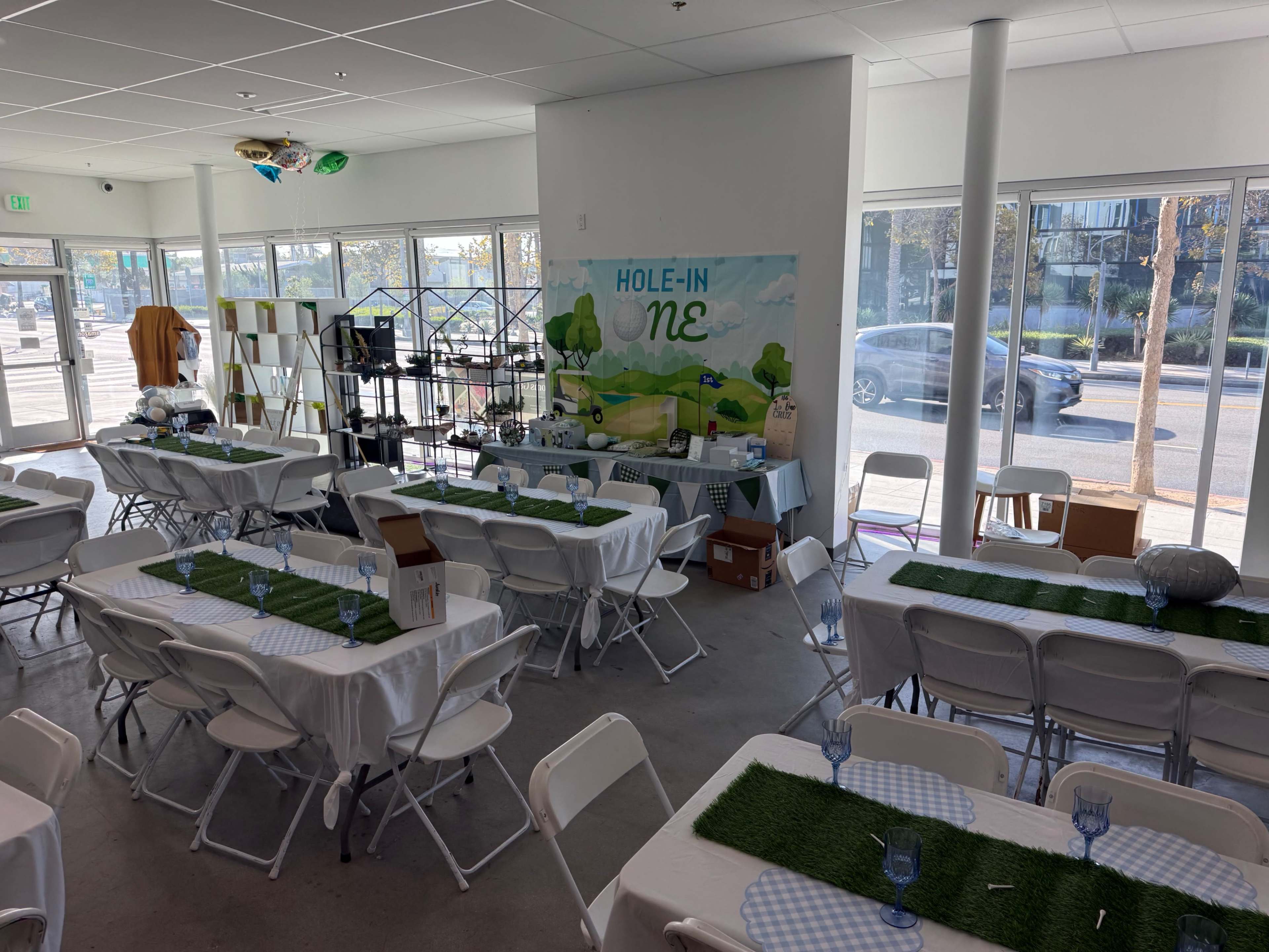 The image shows an indoor event space with tables dressed in white tablecloths and green centerpieces, featuring a mural that reads "HOLE-IN-ONE" on the wall.