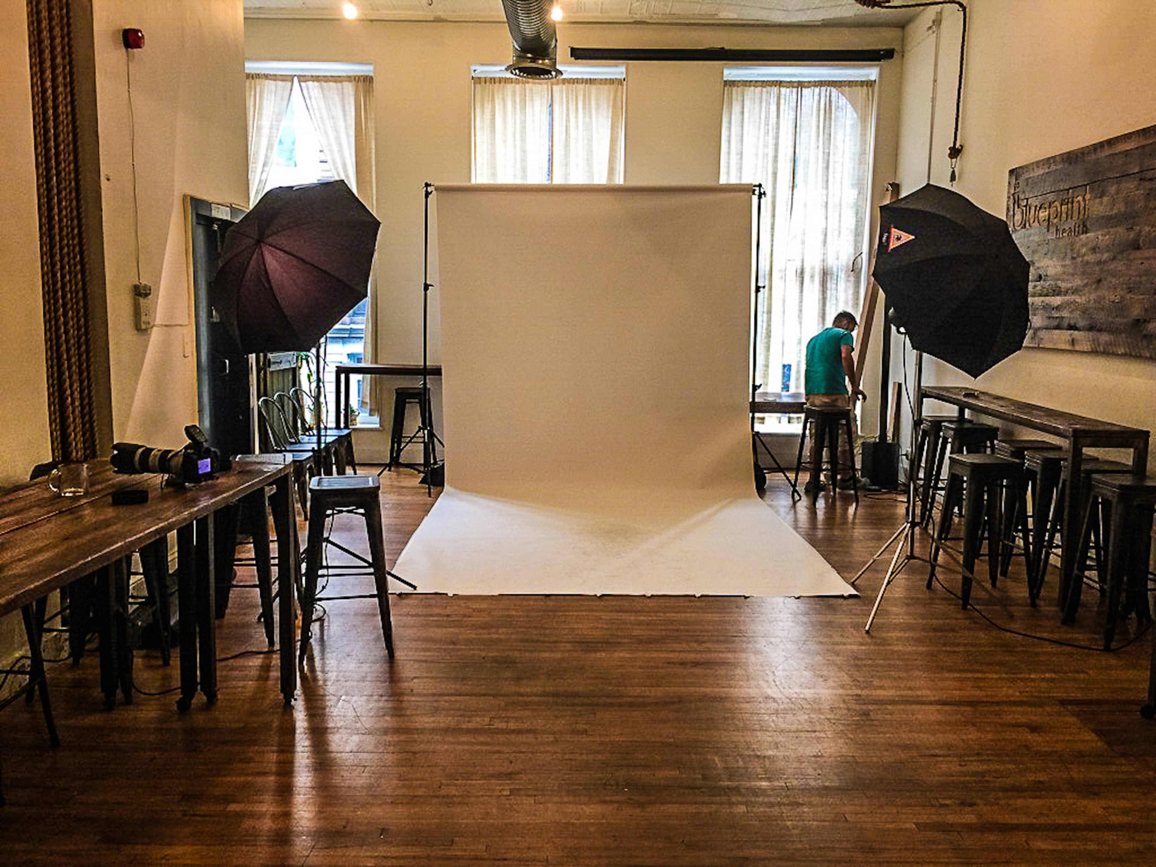 A photography studio setup features a white backdrop, lighting equipment on stands, and a person arranging items in a room with wooden tables and chairs.