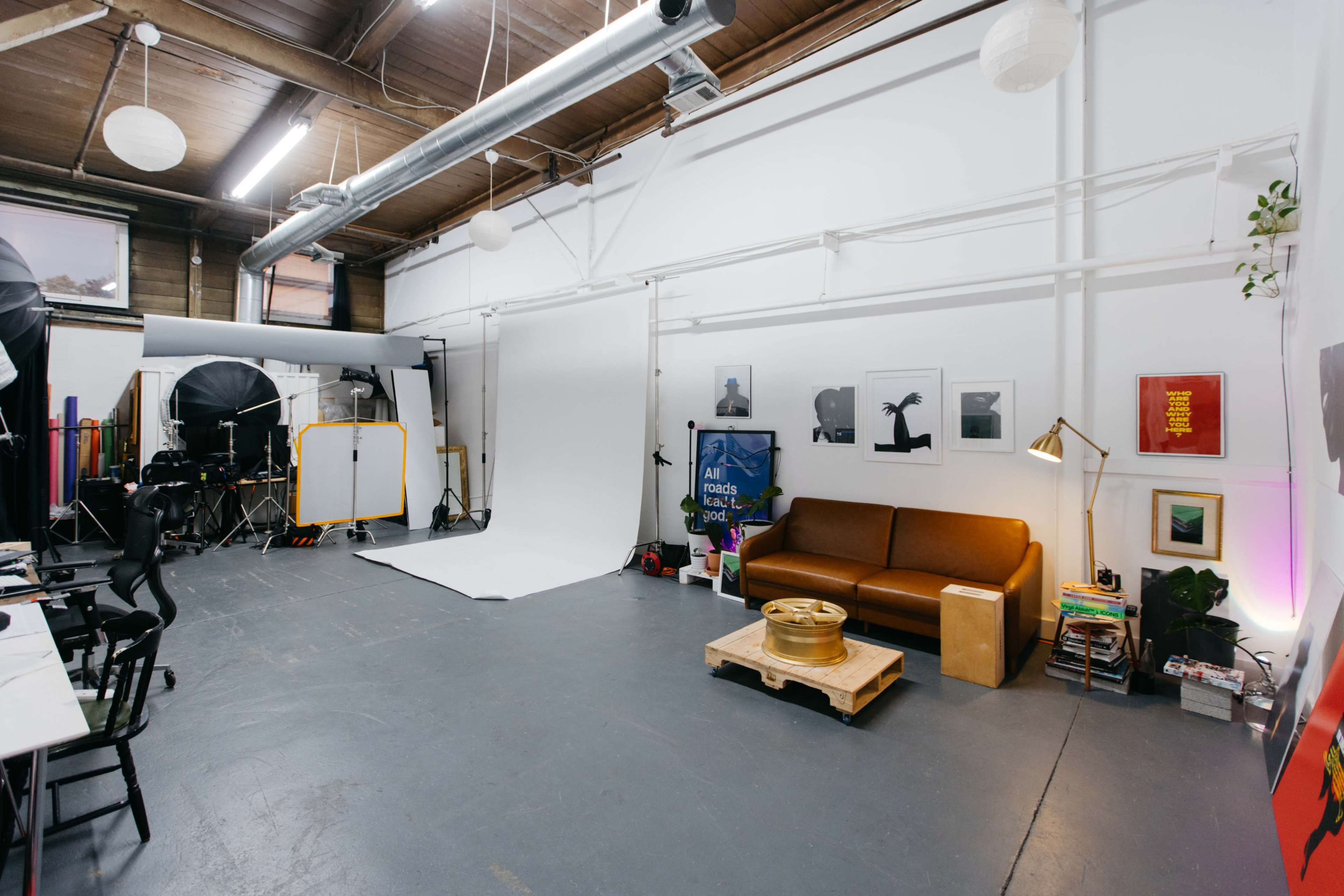 The image shows a spacious photography studio featuring a brown sofa, a backdrop setup, various lighting equipment, and artwork on the walls.
