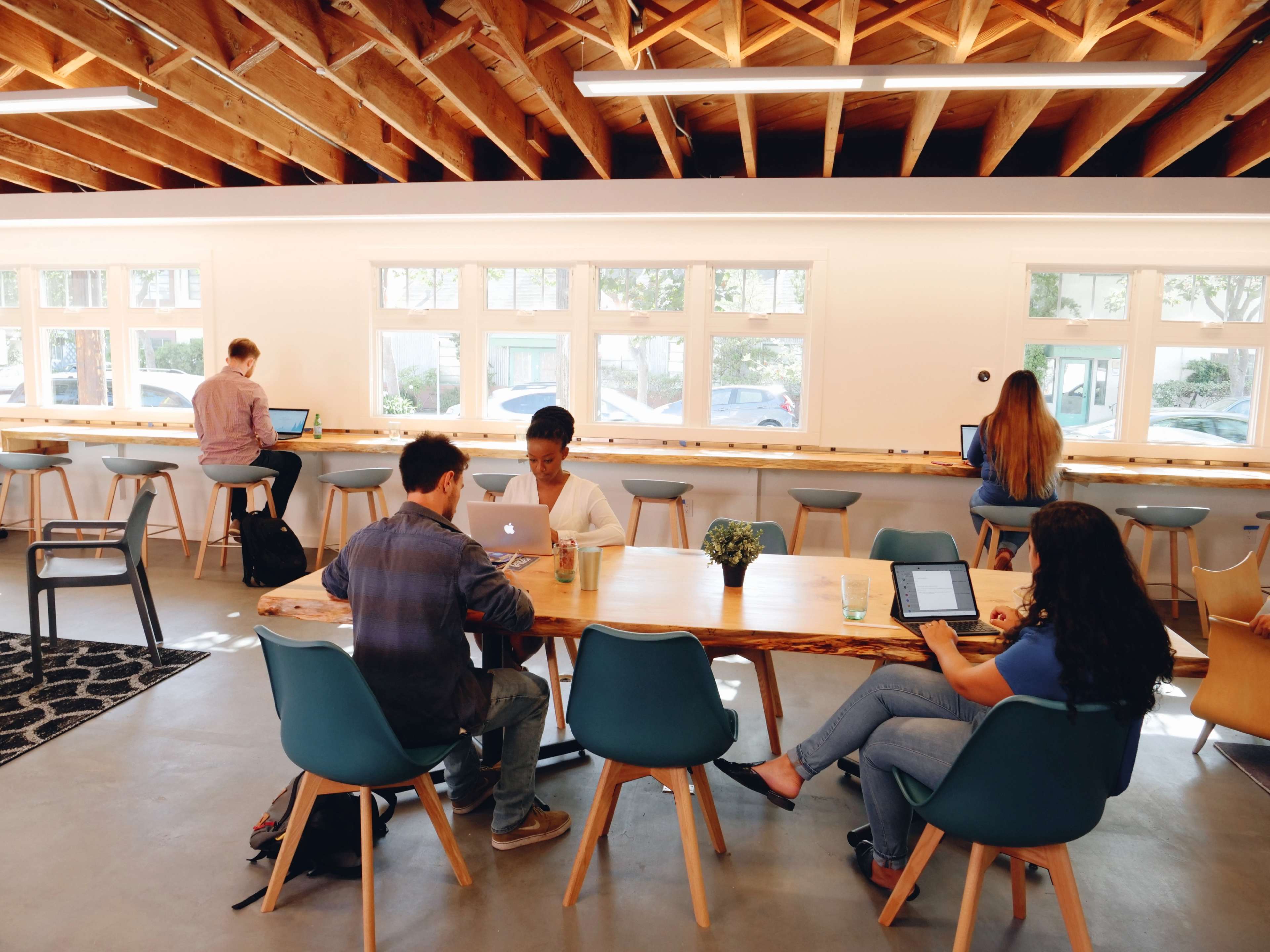 A group of people work on laptops at a communal table in a well-lit, modern workspace with wooden beams and large windows.