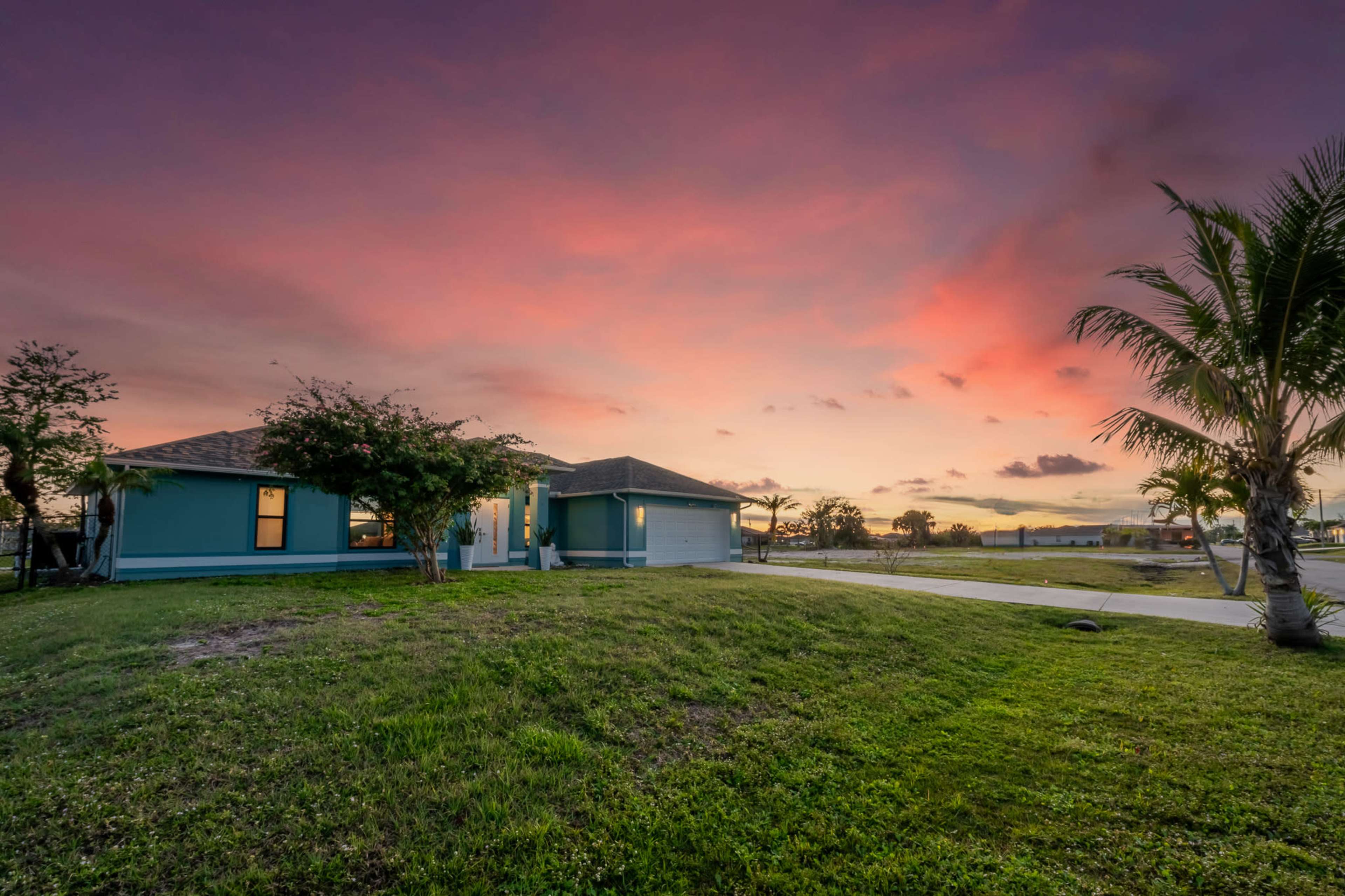 A single-story house with a green exterior is set against a vibrant sunset sky, surrounded by grass and palm trees.