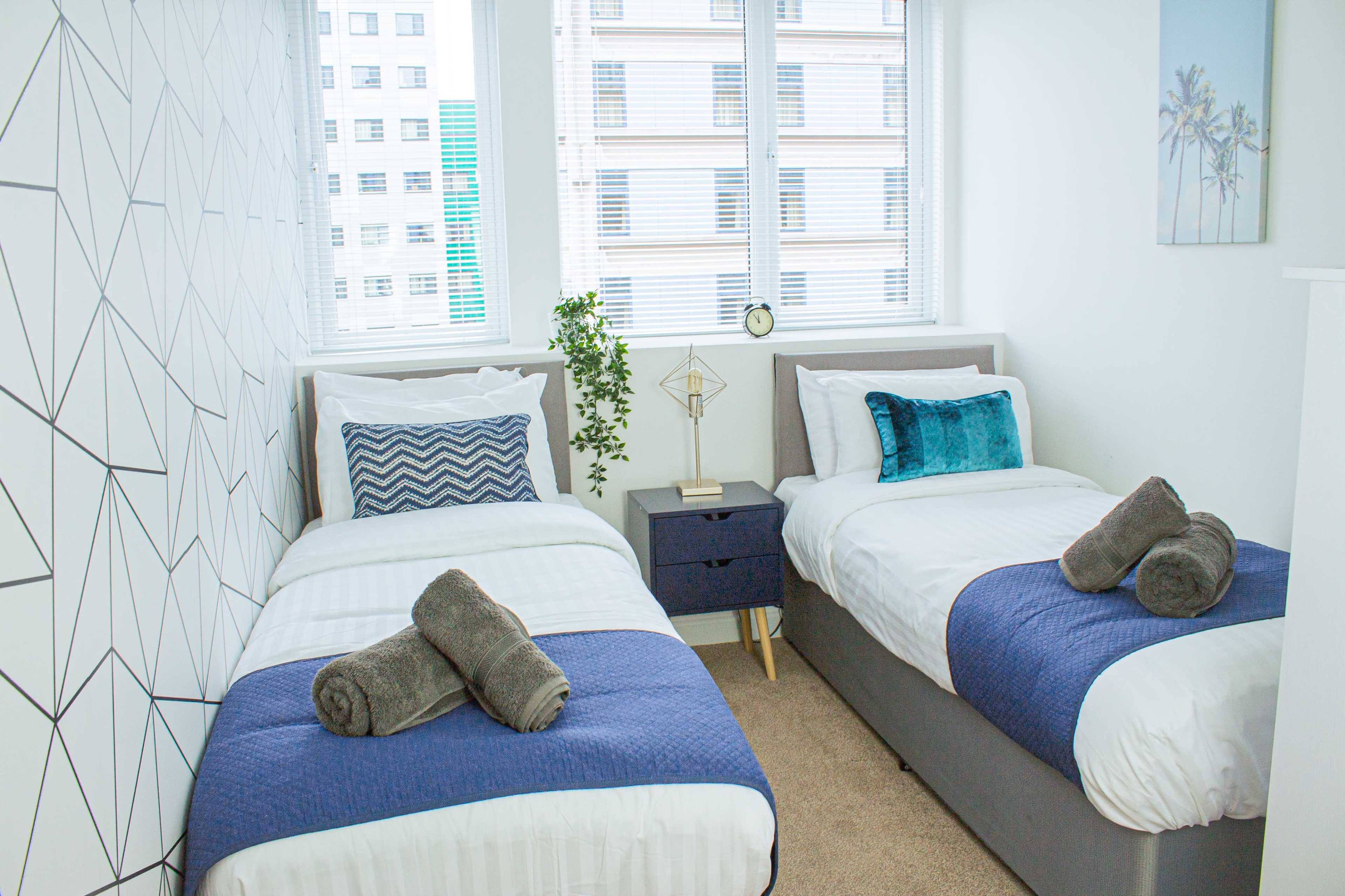 A brightly lit room with two single beds, each adorned with white linens and blue throws, separated by a small nightstand with a decorative item.