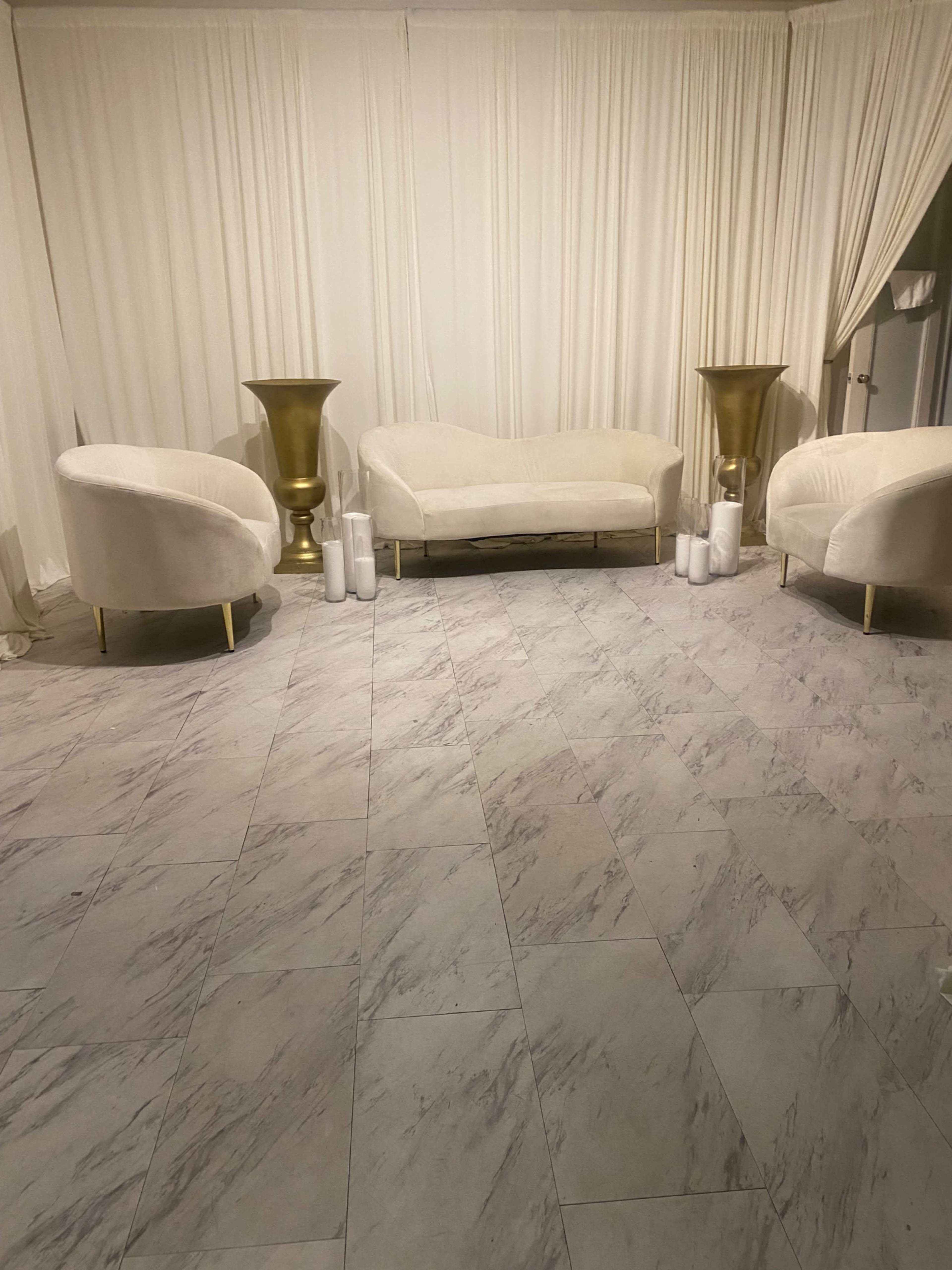 The image shows a minimalist room with two white sofas and two large gold vases, set on a marble-patterned floor with white drapes in the background.