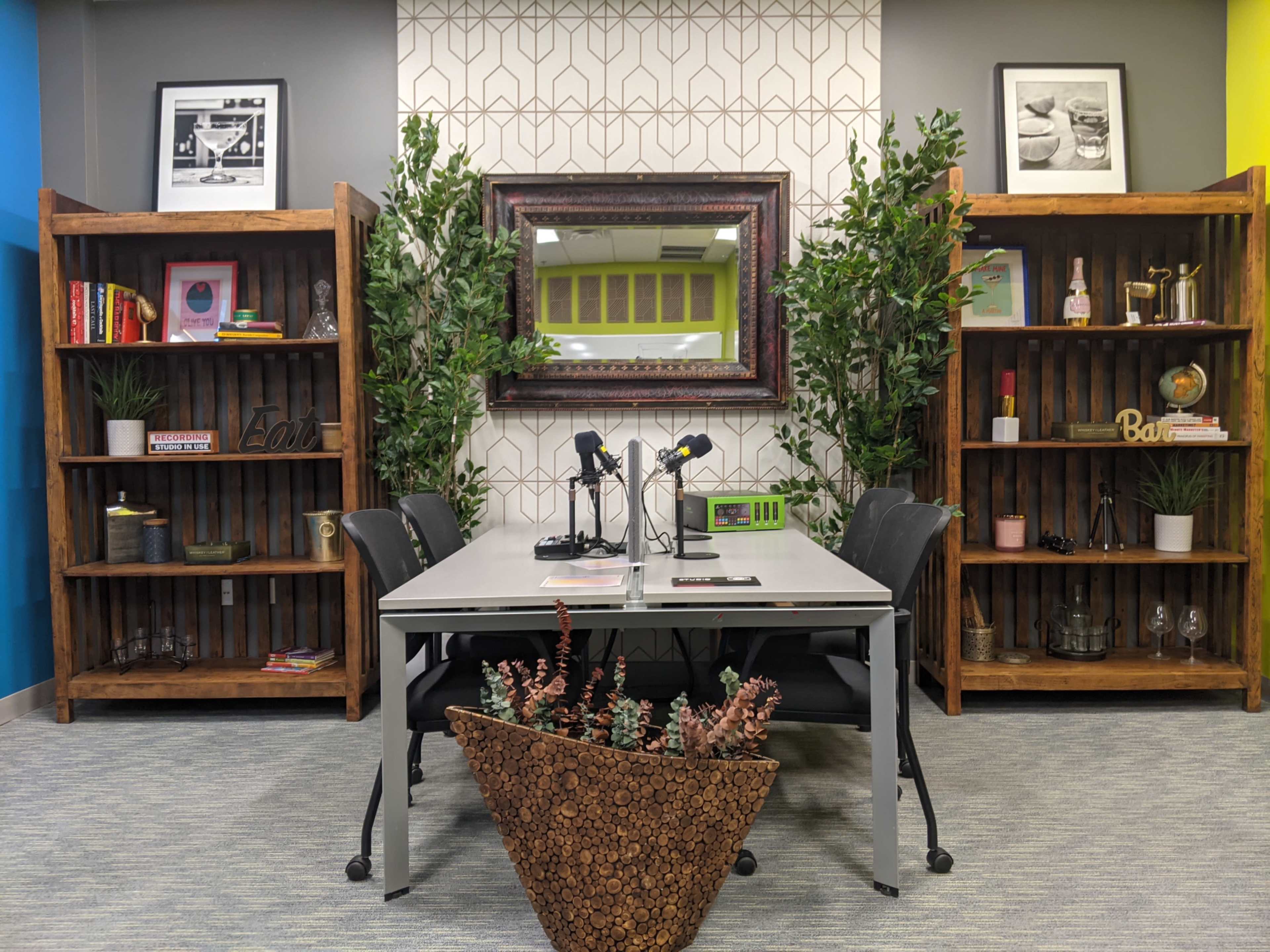 The image shows a modern office space with a rectangular table, two microphones, and shelves filled with books and decor on either side, alongside a large mirror and plant arrangements.