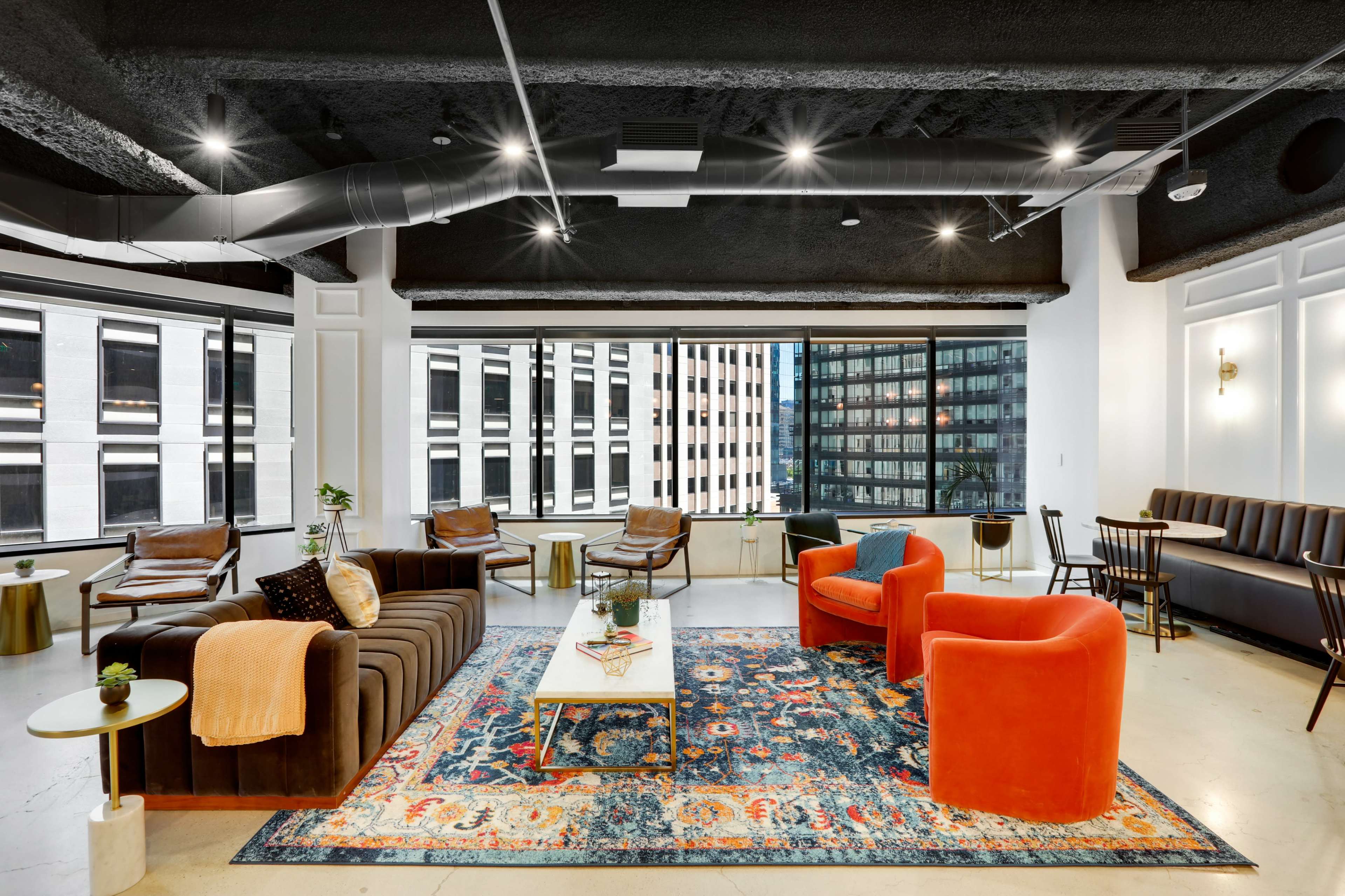 A modern office lounge area with a mix of seating options, including orange armchairs, a dark couch, and a patterned rug under a bright, spacious window.