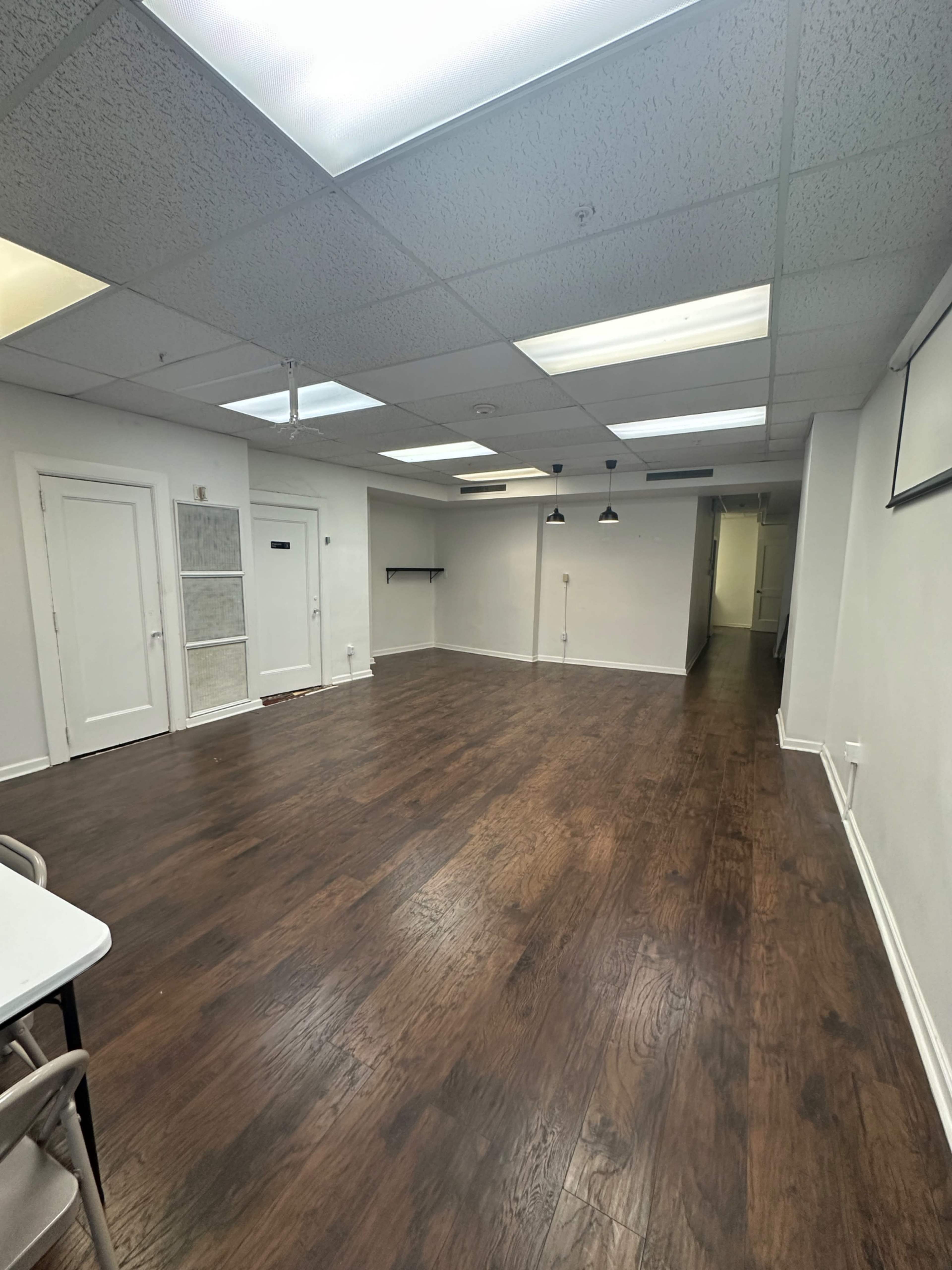 The image shows a spacious, empty room with wooden flooring, overhead lights, and white walls.