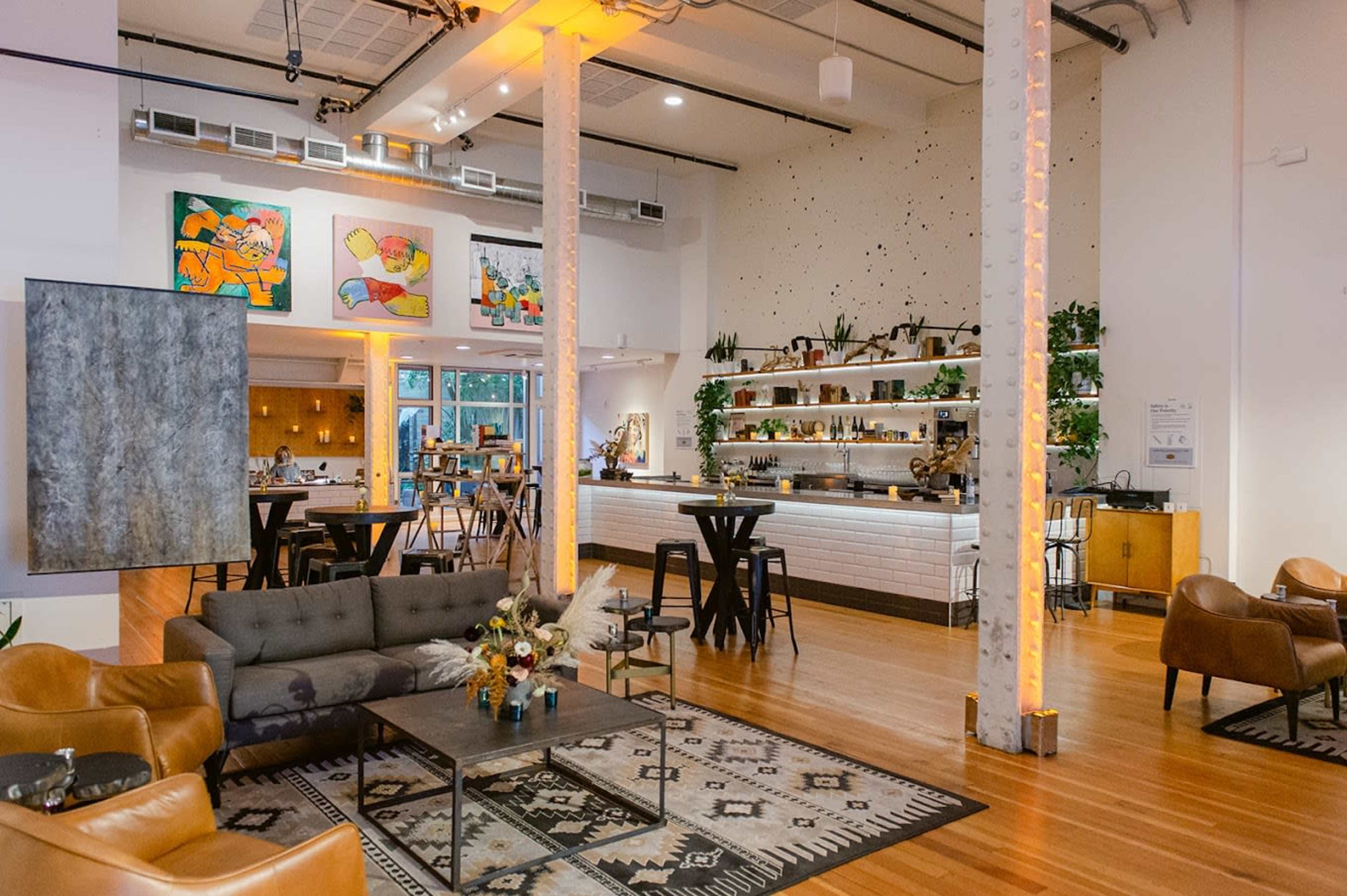 The image shows a modern, open-concept space featuring a lounge area with seating, a bar with plants, and colorful artwork on the walls.