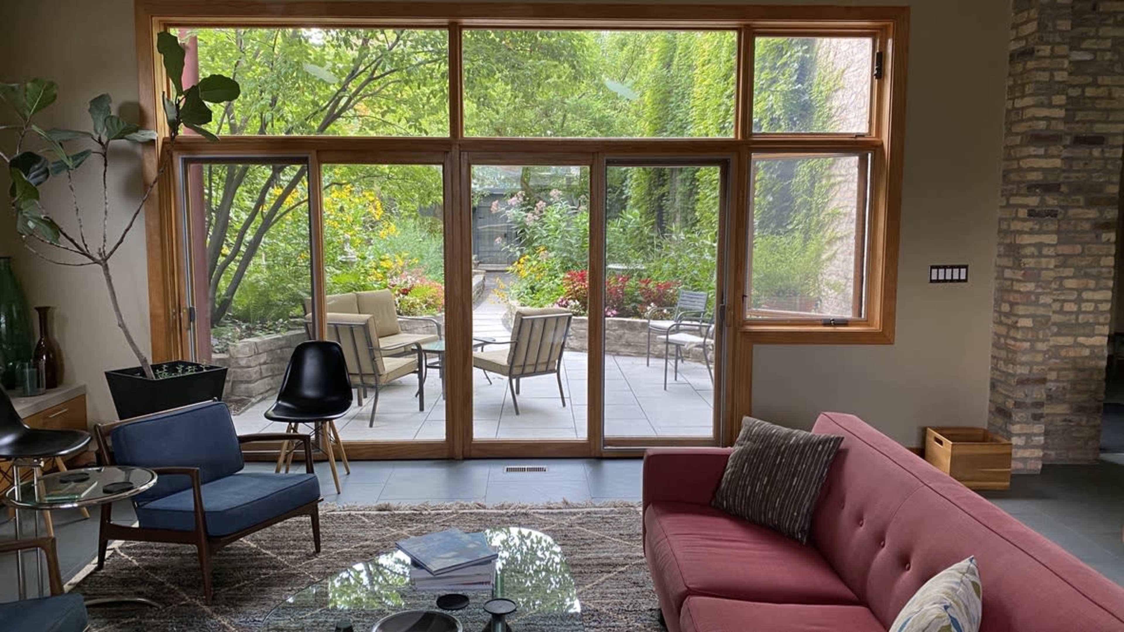 A modern living room features a red couch and chairs, with large glass doors opening to a garden patio filled with greenery and outdoor furniture.