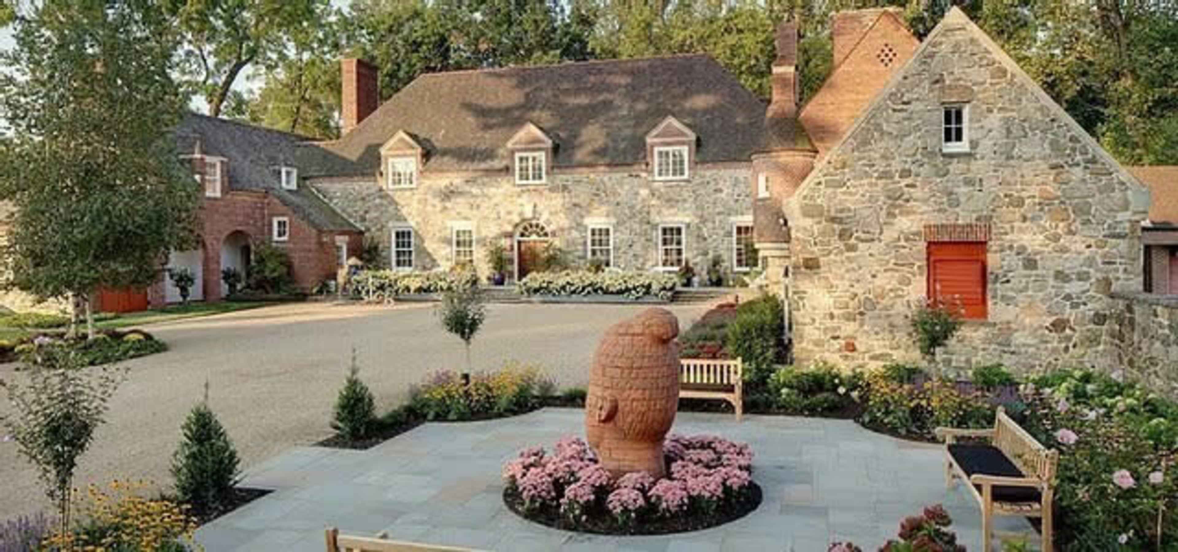 A landscaped courtyard featuring a stone house, a smaller stone structure, a central sculpture of a beehive, and surrounding flower beds with benches.