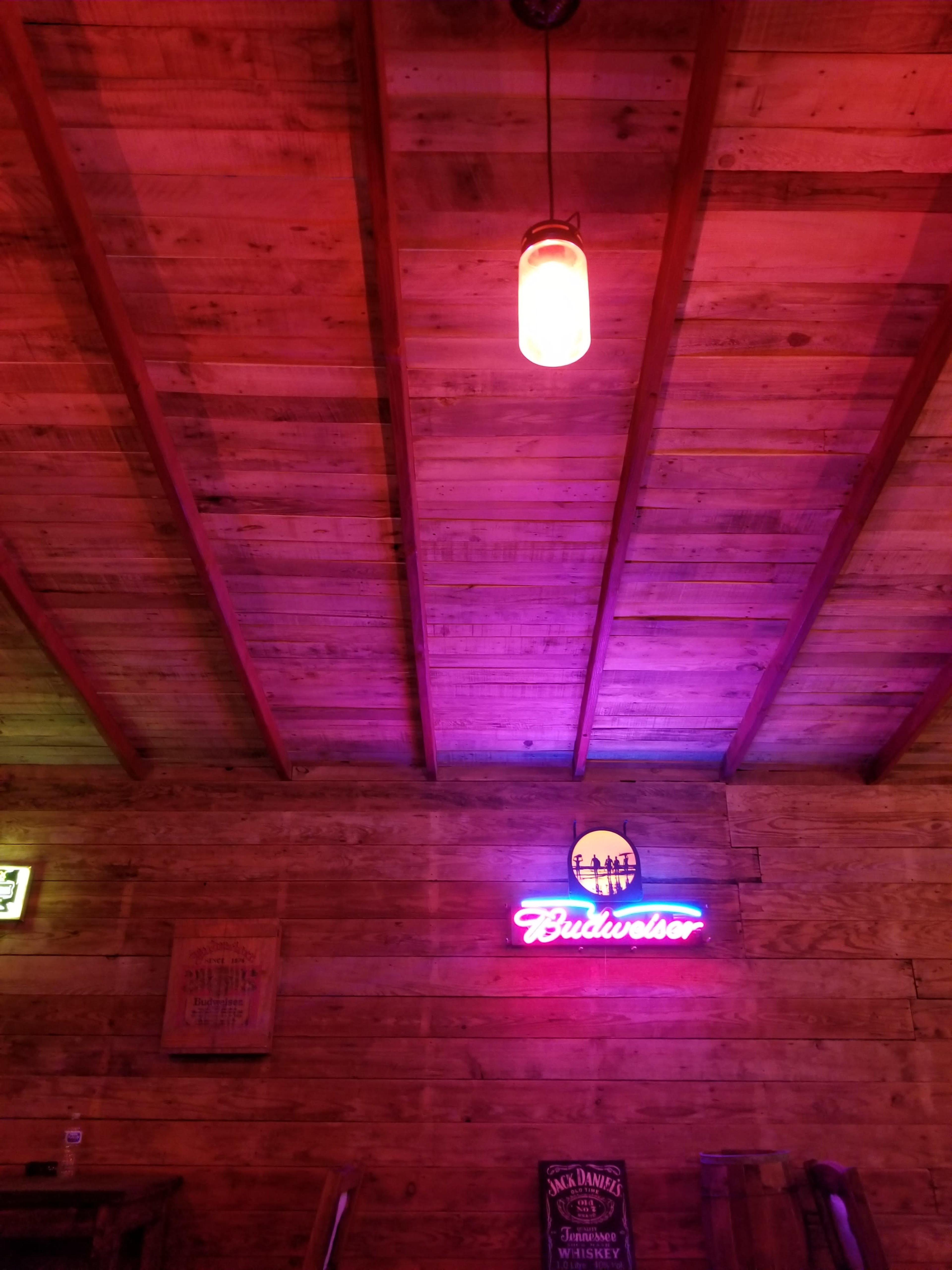 A wooden interior with pink and purple lighting, featuring a Budweiser neon sign and a hanging light fixture.