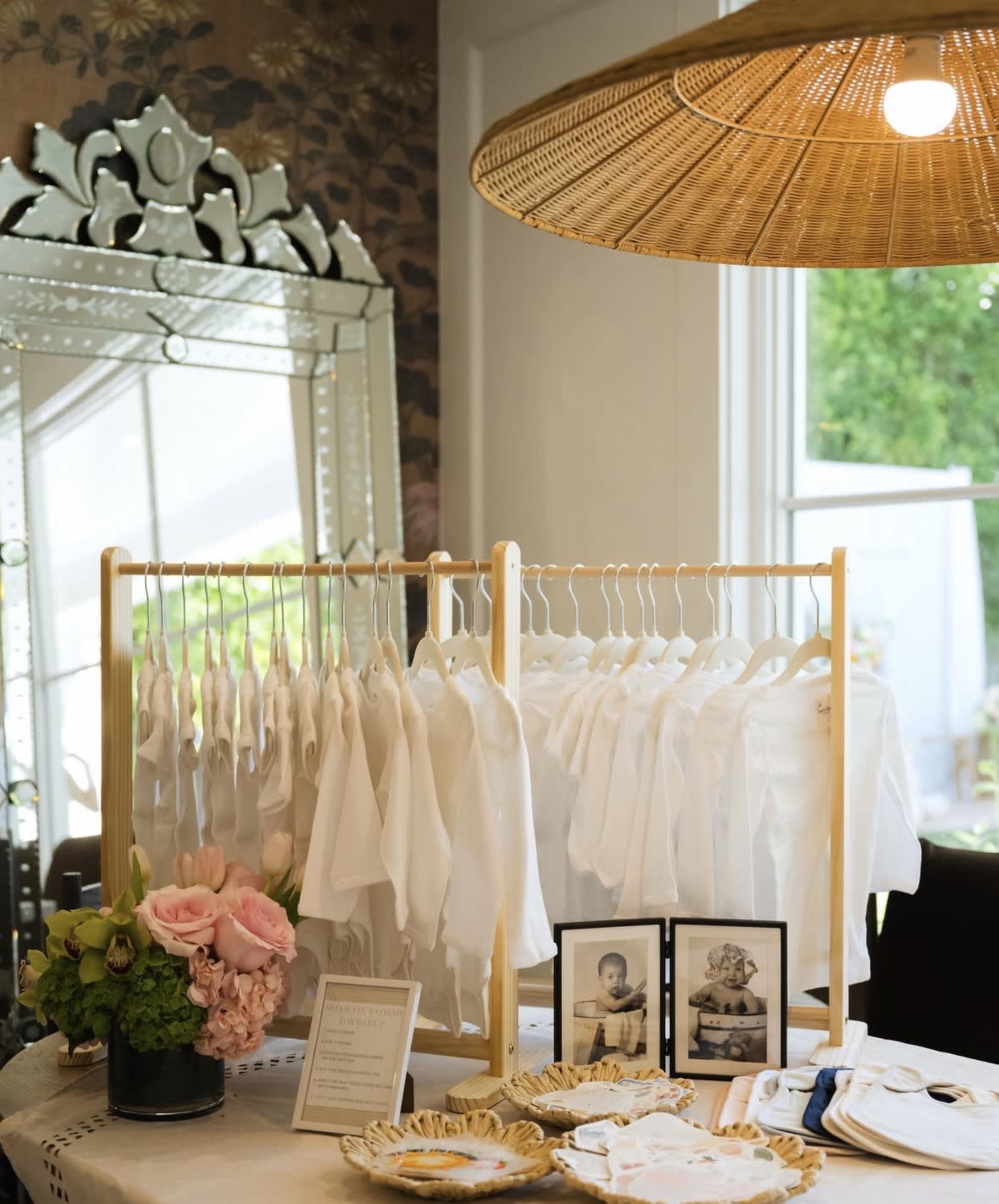 A display featuring white clothing on hangers, surrounded by floral arrangements and framed photographs, is set up near a large mirror and an overhead pendant light.