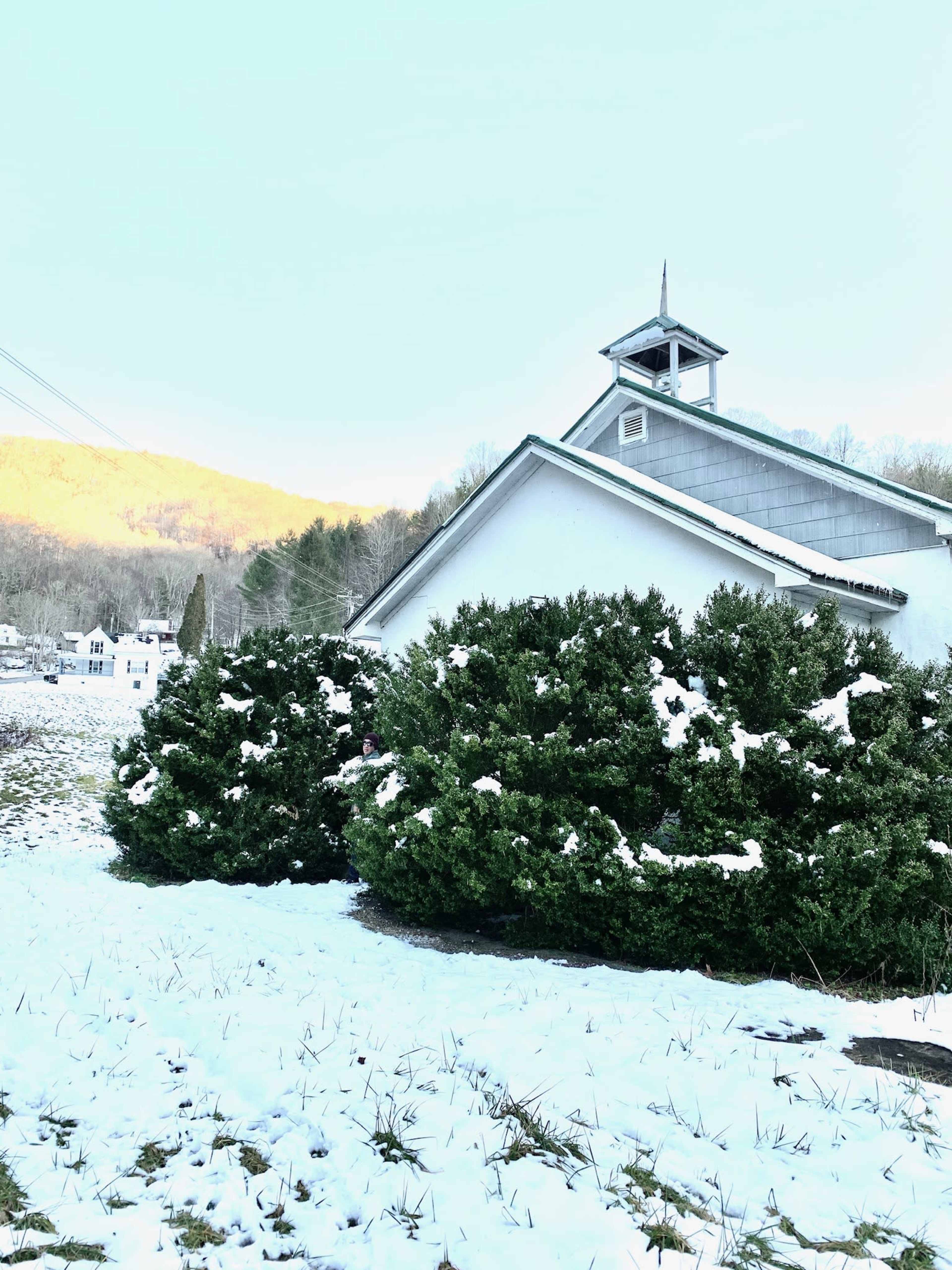 A snow-covered church stands beside a row of bushy green shrubs, with a mountainside visible in the background.