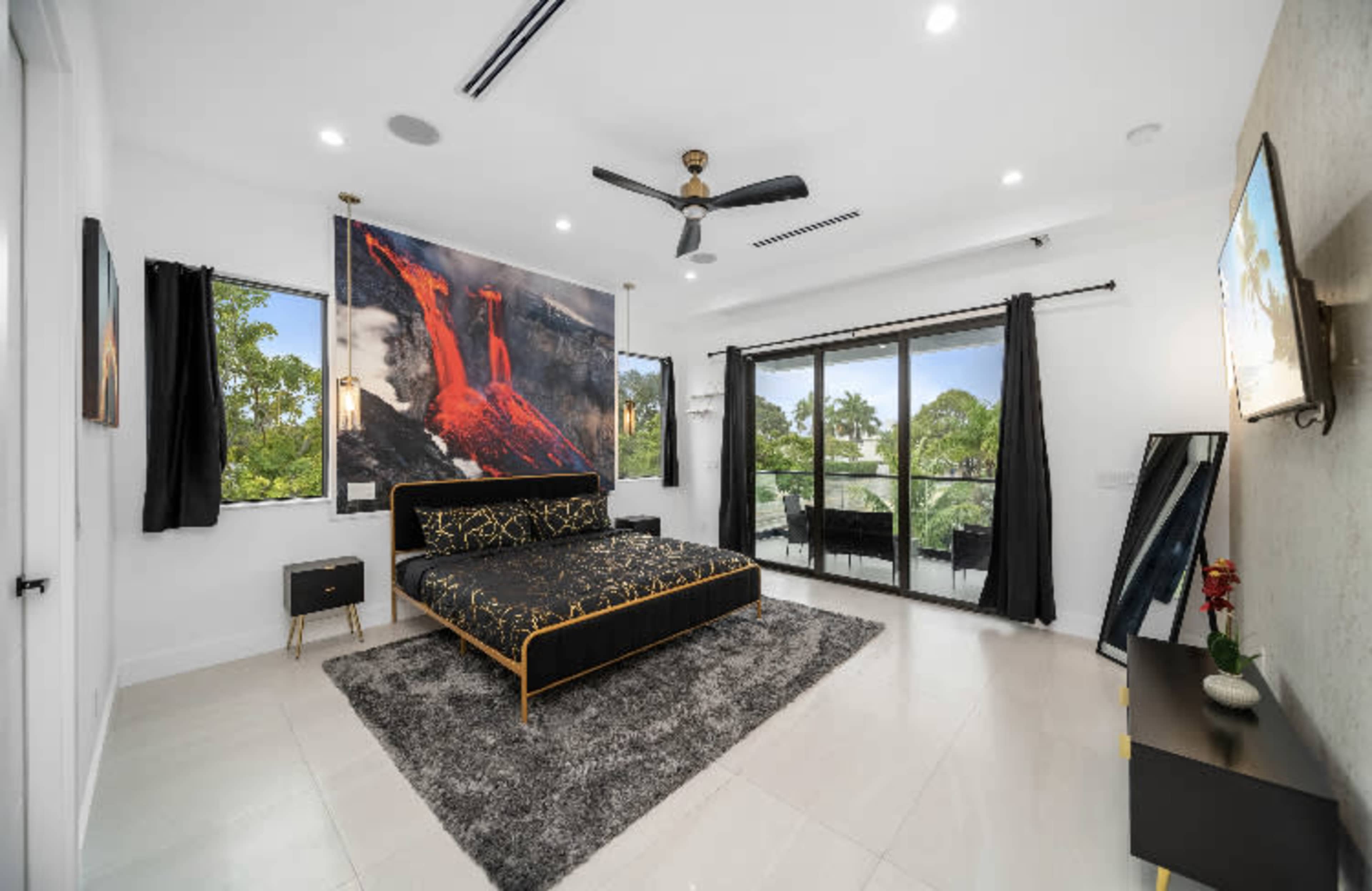 A modern bedroom features a black bed with a large mural, a ceiling fan, and glass doors leading to a balcony.