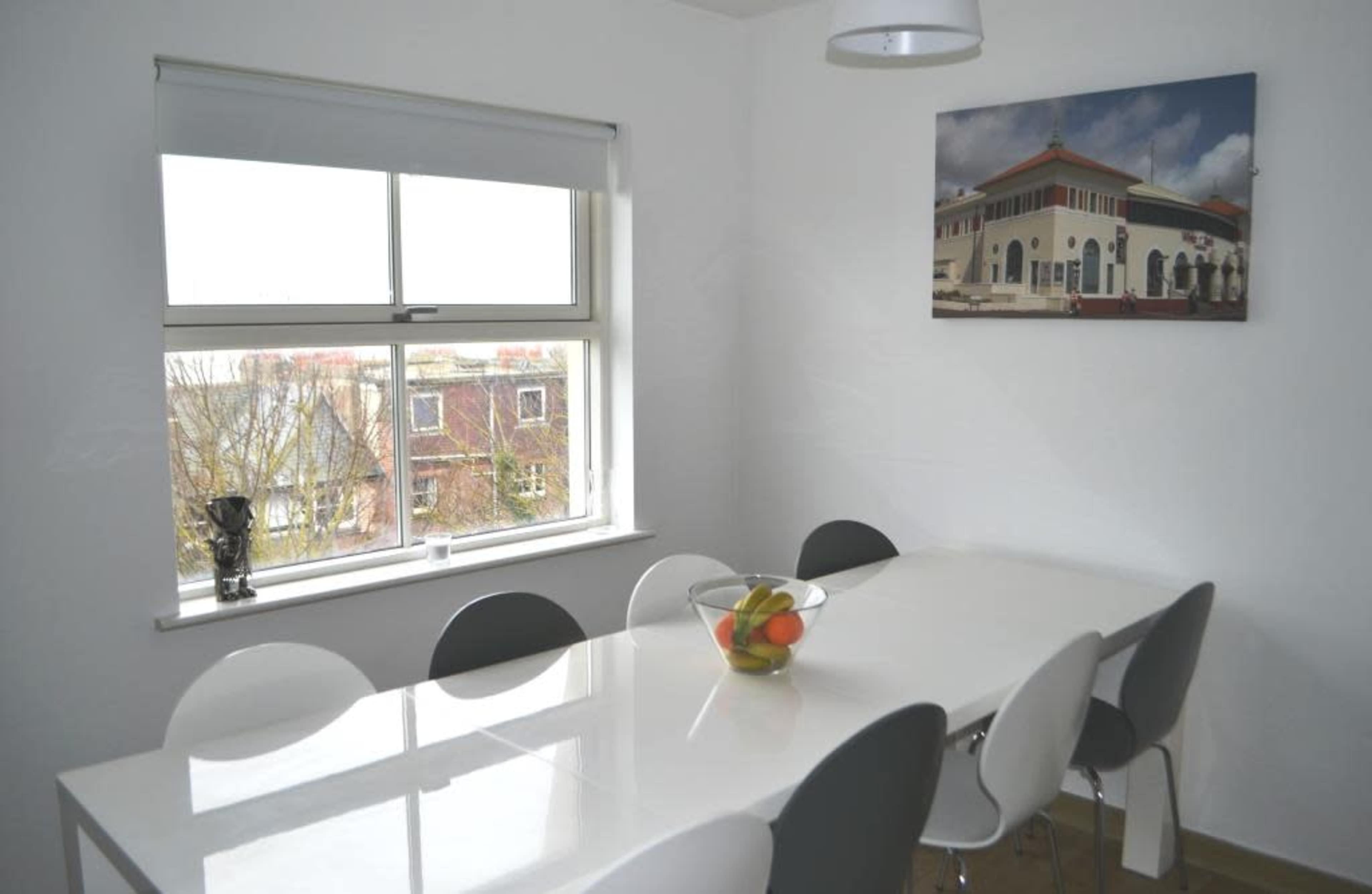 A modern dining room features a large white table surrounded by gray and white chairs, with a bowl of fruit on the table and a window showing a view of nearby buildings.